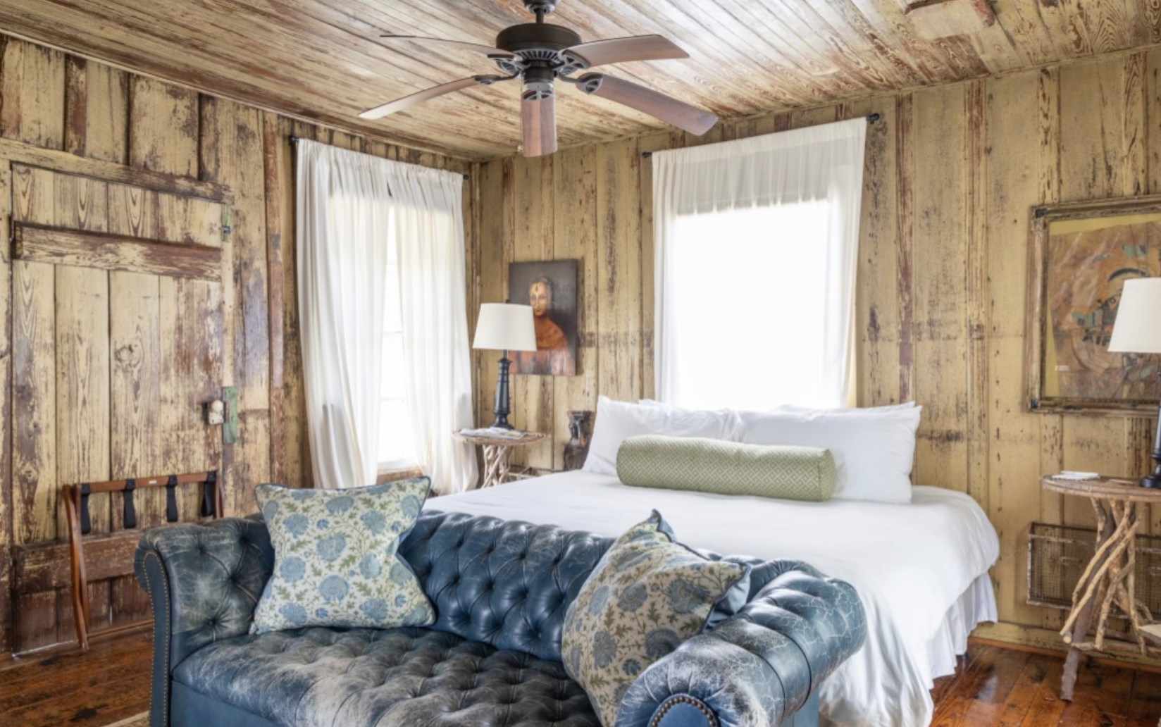 A rustic bedroom with wood-paneled walls and ceiling, featuring a queen-sized bed with white linens and assorted pillows. There is a blue tufted leather sofa with patterned pillows in front of the bed, and two windows with white curtains. The room has a ceiling fan, and side tables with lamps and artwork.
