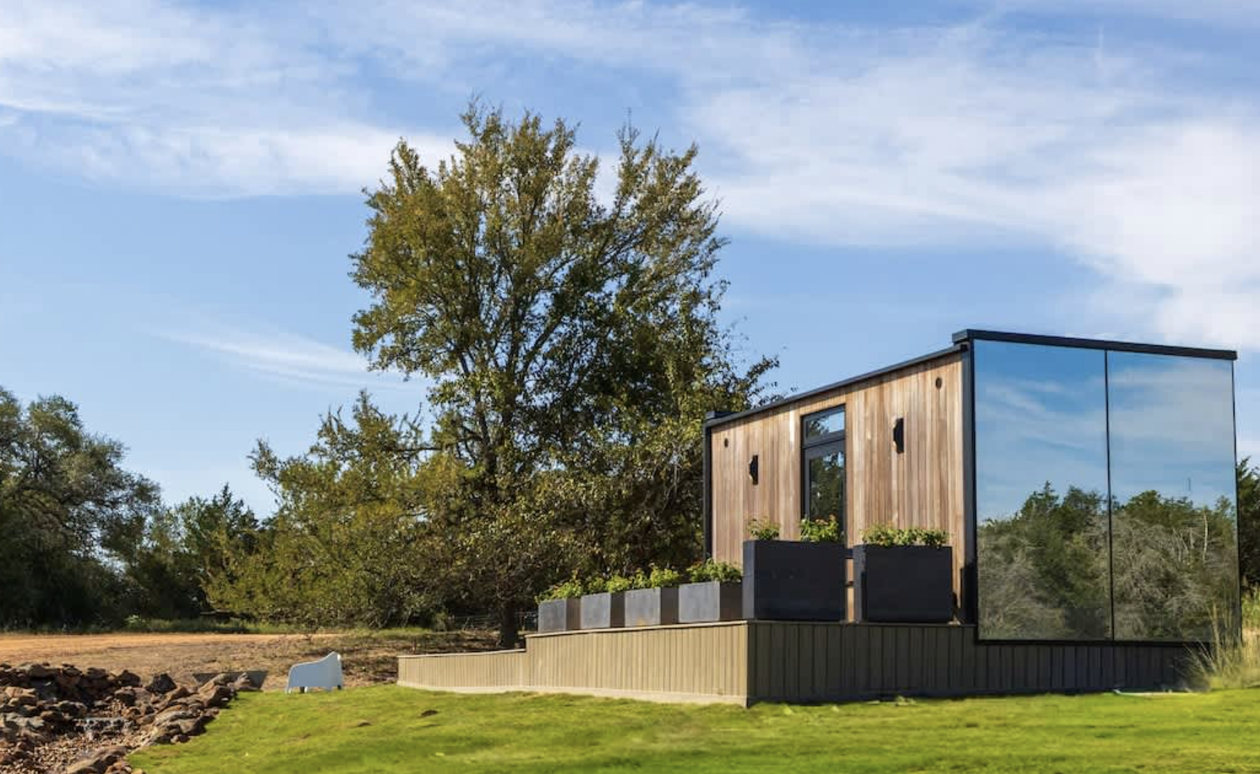 Modern tiny house with wooden exterior and large mirror-like glass wall, surrounded by green grass and trees, under a partly cloudy sky.