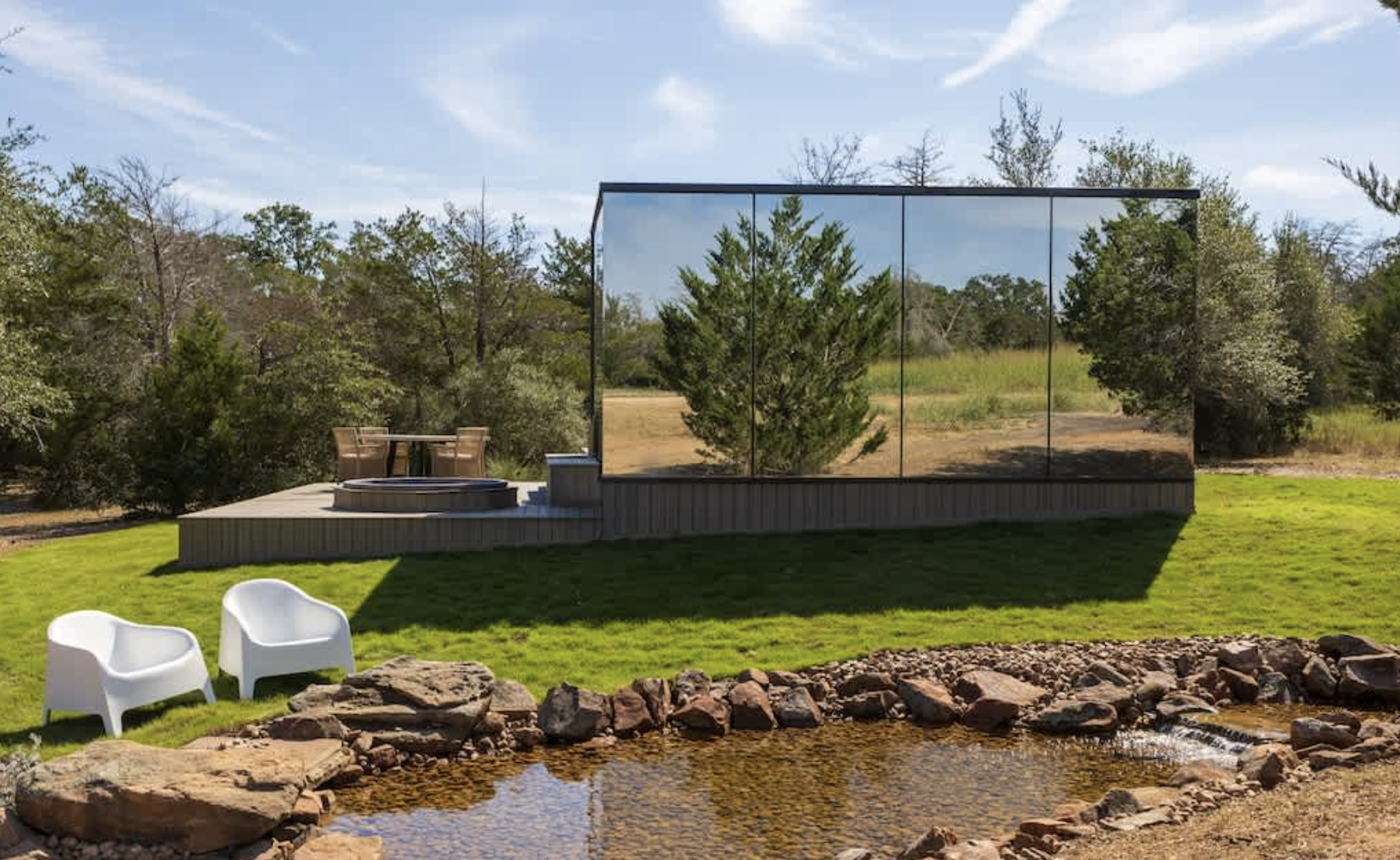 A backyard scene with a small pond and two white chairs in the foreground, a grassy area, a deck with a hot tub, and a tall glass wall reflecting trees and sky in the background.