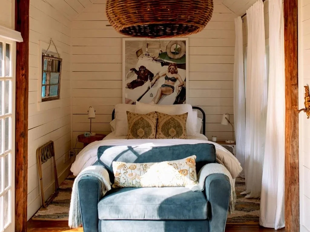 A cozy bedroom with white wood-paneled walls, featuring a bed with white bedding, decorative pillows, and a framed photo of people on a boat. A small wooden side table with a lamp, an armchair with a patterned pillow, and curtains covering windows add to the inviting atmosphere.