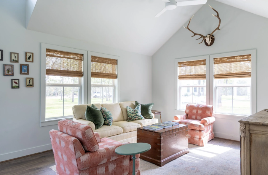 Bright living room with white walls, four large windows with bamboo shades, a cream sofa with green and patterned pillows, two pink plaid armchairs, a wooden coffee table with magazines, a small round side table, a rustic wooden cabinet, and a mounted deer antler on the wall.