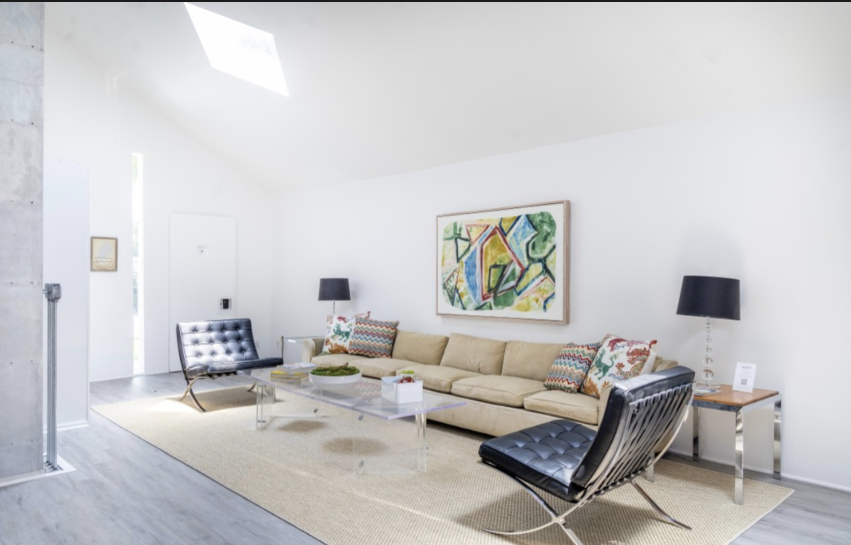 Living room with cream sofa, two black leather chairs, a glass coffee table, abstract art on white walls, and black lamps.