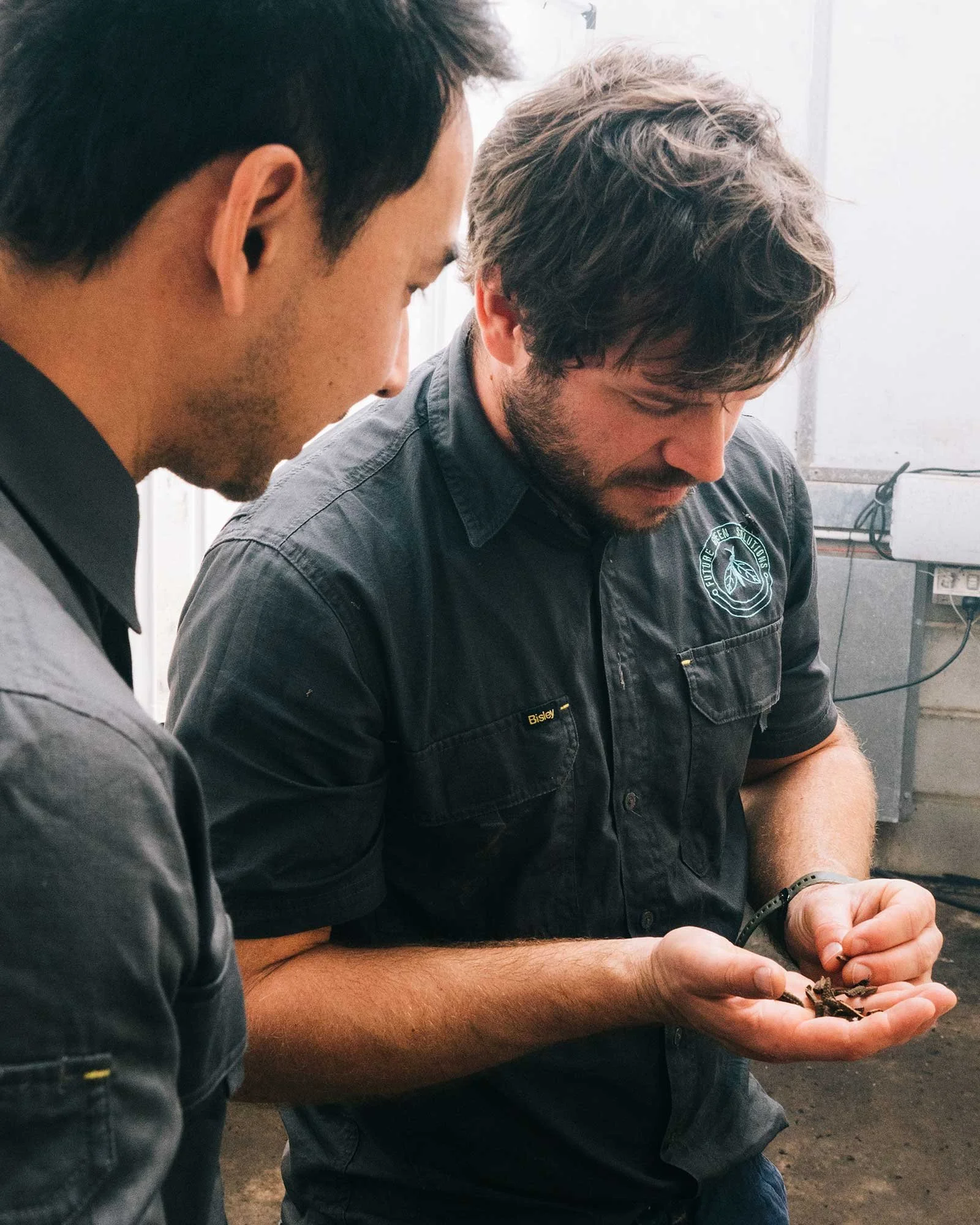 Two men are closely examining objects in their hands, with one wearing a black shirt with a patch on the sleeve and a badge that reads "Bisley." They are indoors, possibly in a workshop or laboratory.