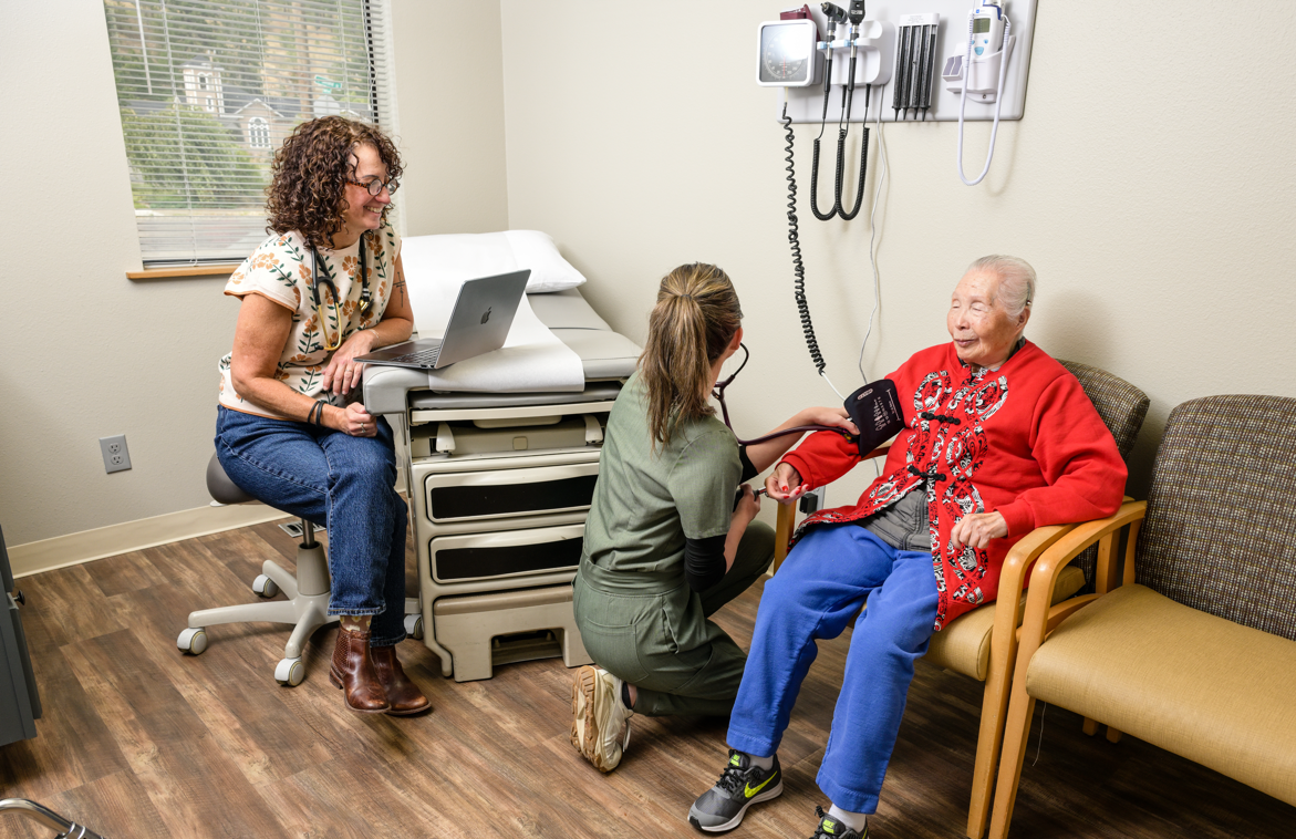 Hayden takes blood pressure on a smiling patient as Bobbi watches with a smile in an exam room