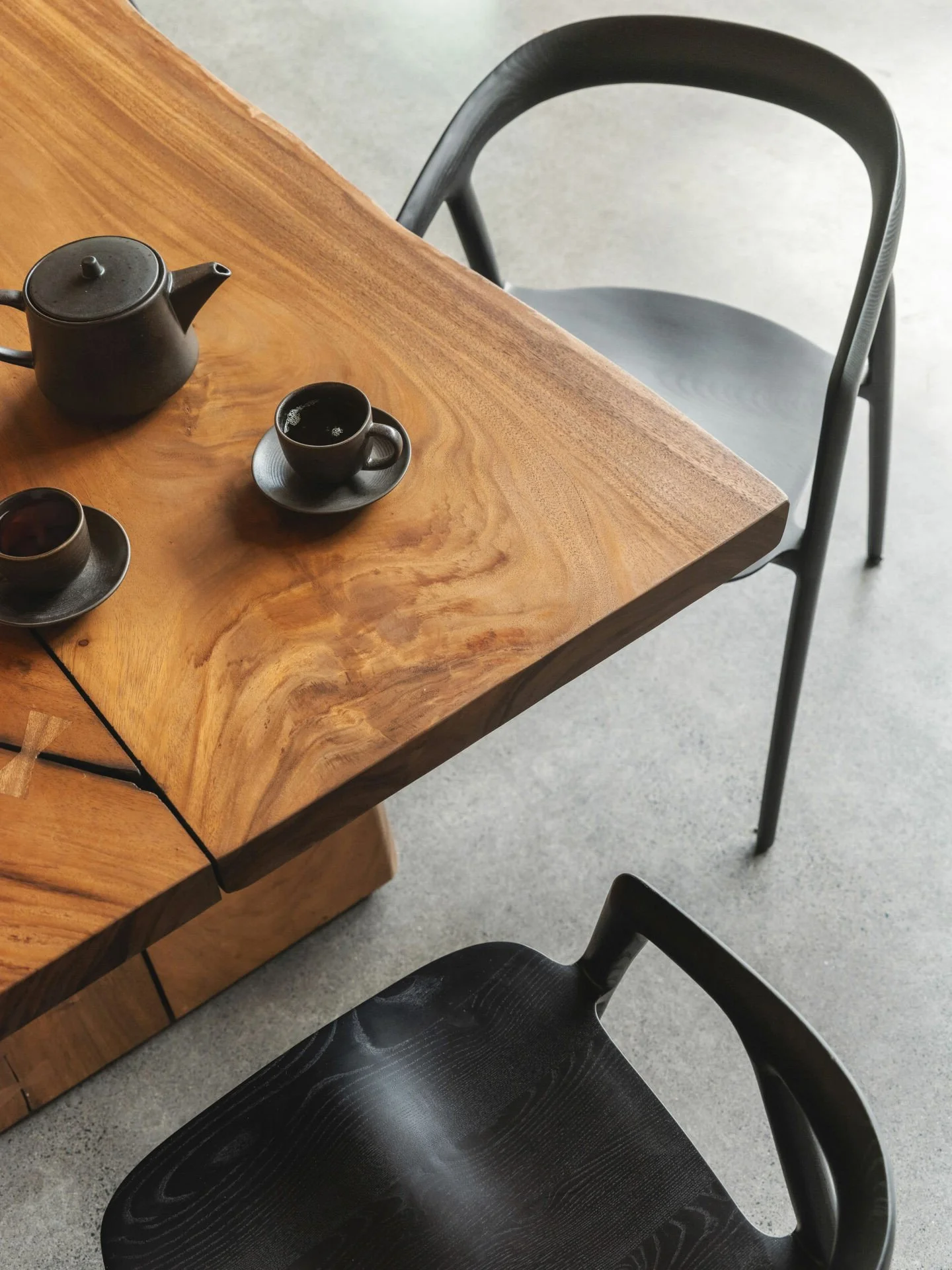 A wooden table with a dark teapot and two cups on matching saucers, two black chairs, and a concrete floor.
