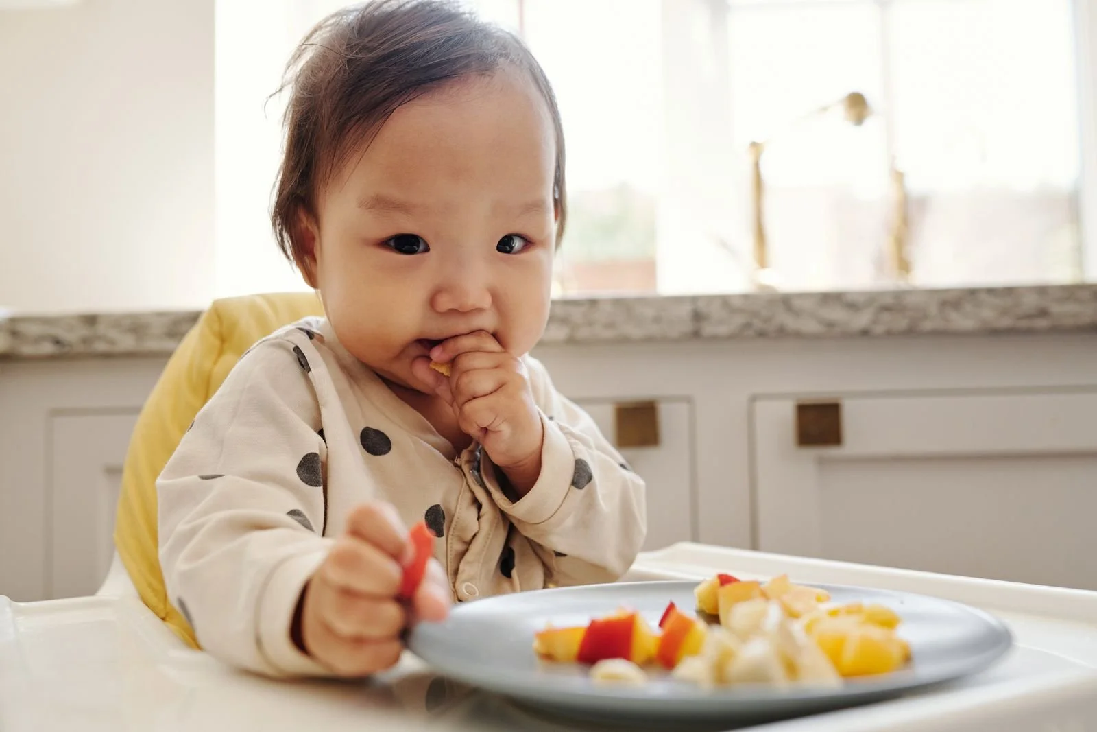 Toddler in a high chair eating bite-sized fruit from a plate on the tray
