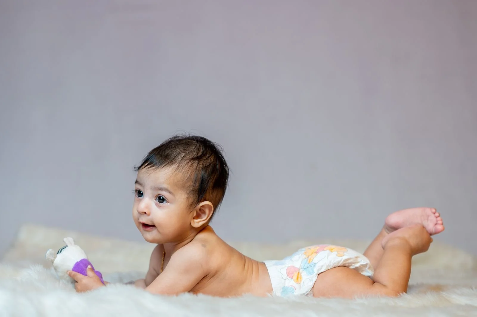 Smiling baby lying on their tummy on a soft blanket, propped on their forearms and holding a toy