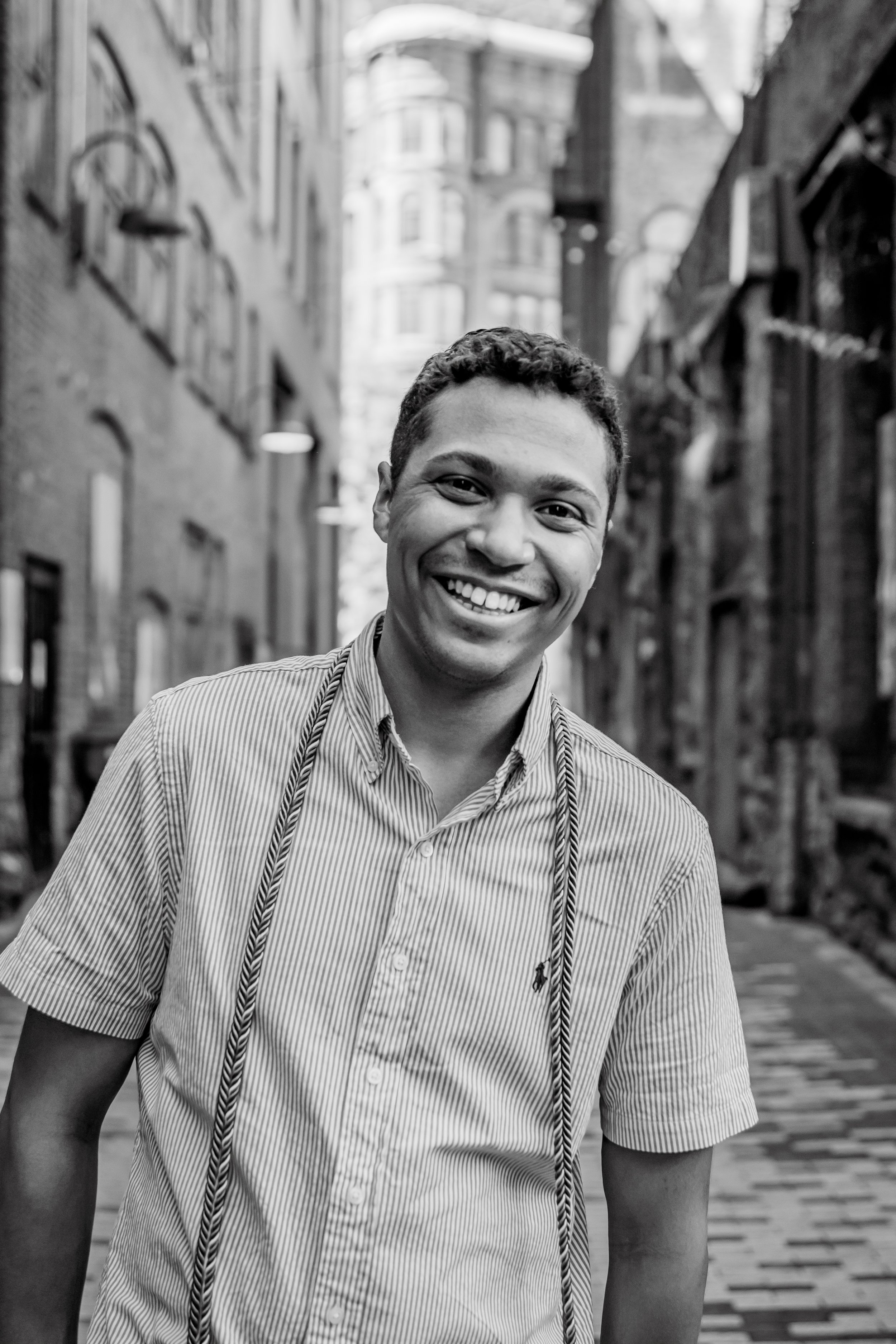A young man smiling towards the camera pictured in black and white in the middle of an alleyway with buildings.