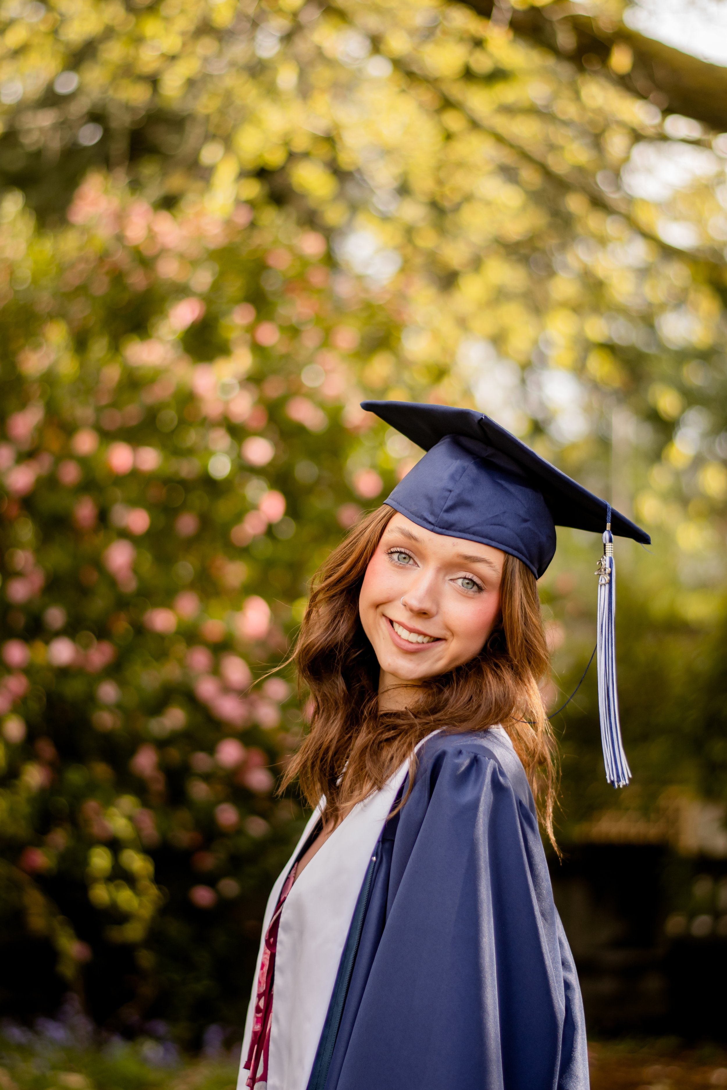 A young woman smiling in her graduation regalia in front of flowers and greenery.