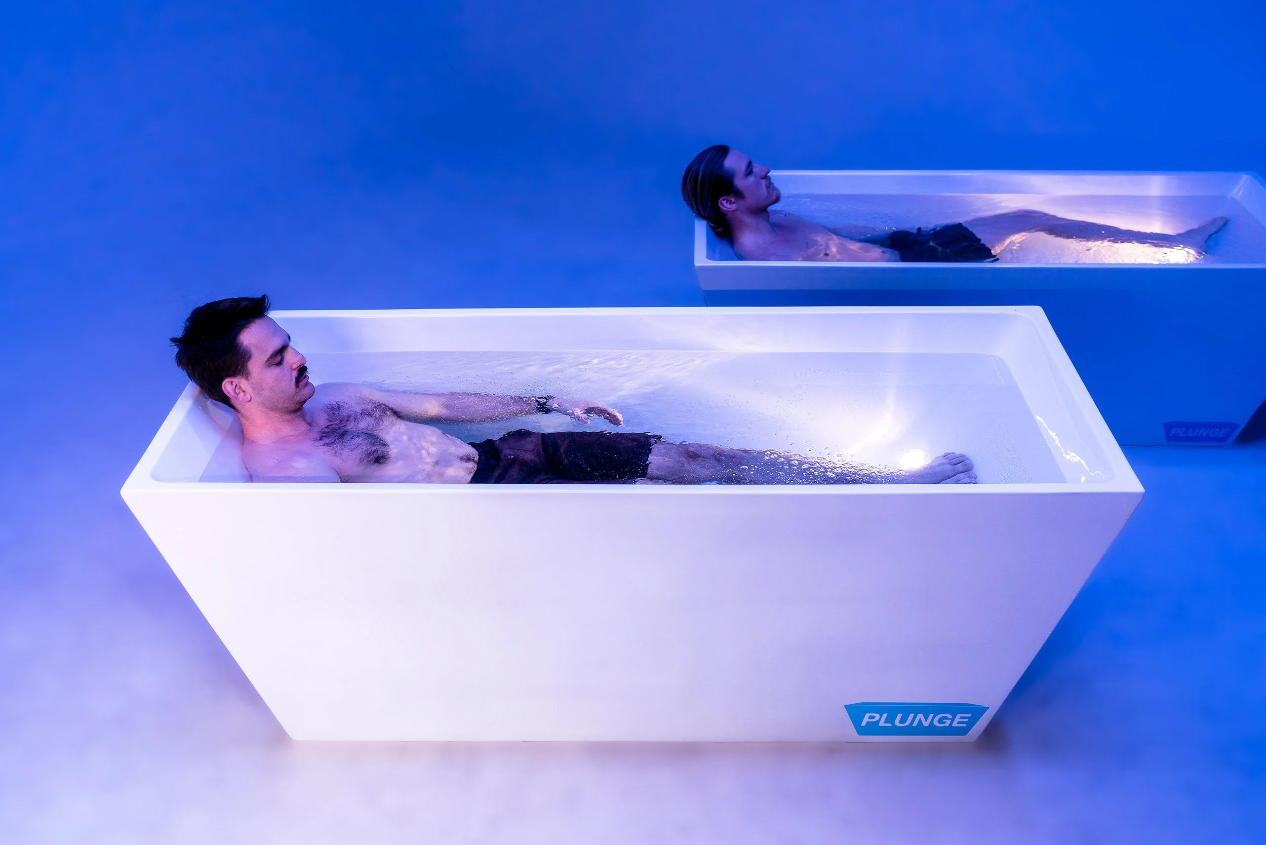Two men lying in separate white rectangular floating tanks filled with water, with a blue background and soft lighting.