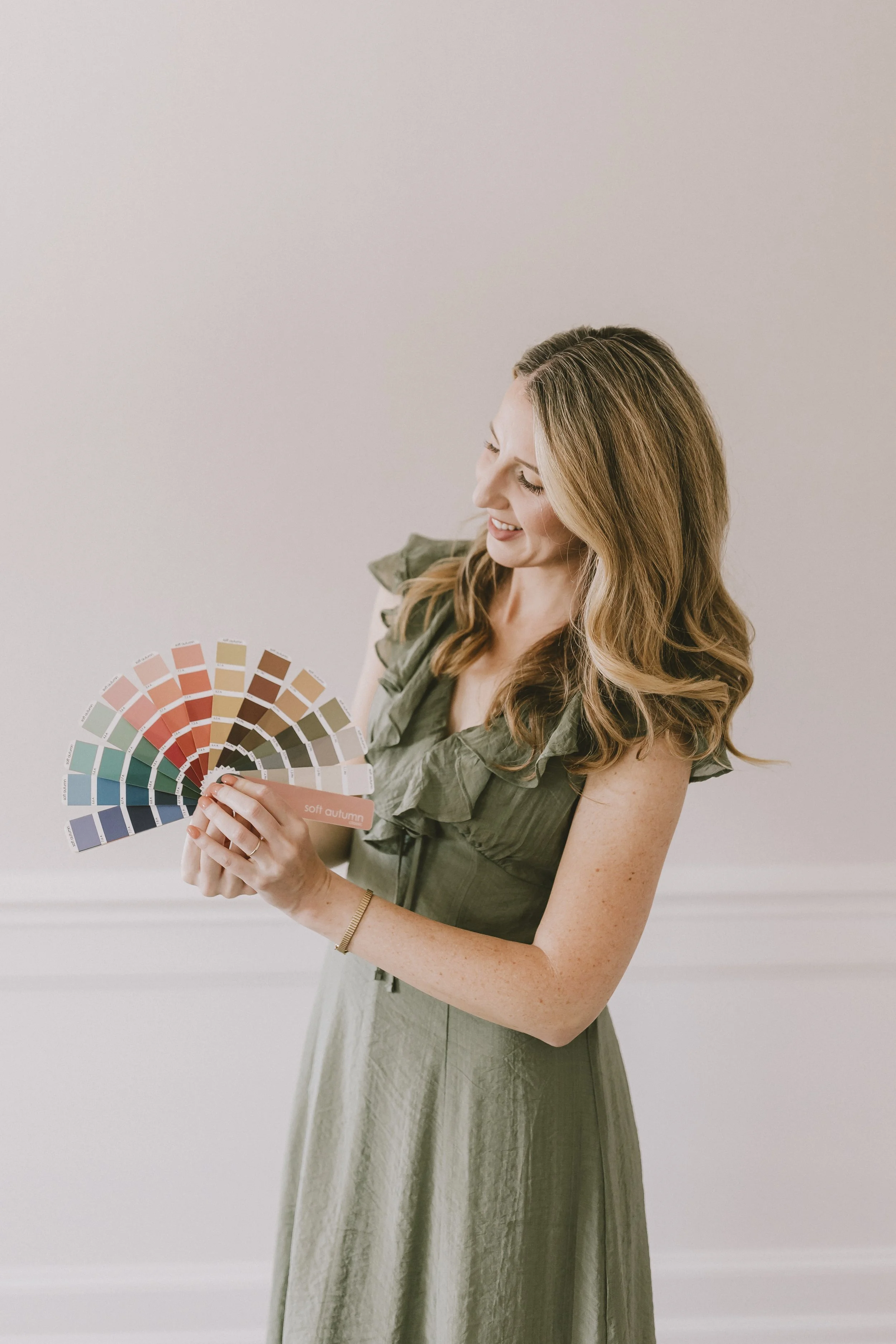 A woman with long wavy hair wearing a green dress and gold bracelet, smiling while holding a color swatch fan labeled 'soft autumn' in front of a plain light-colored wall.