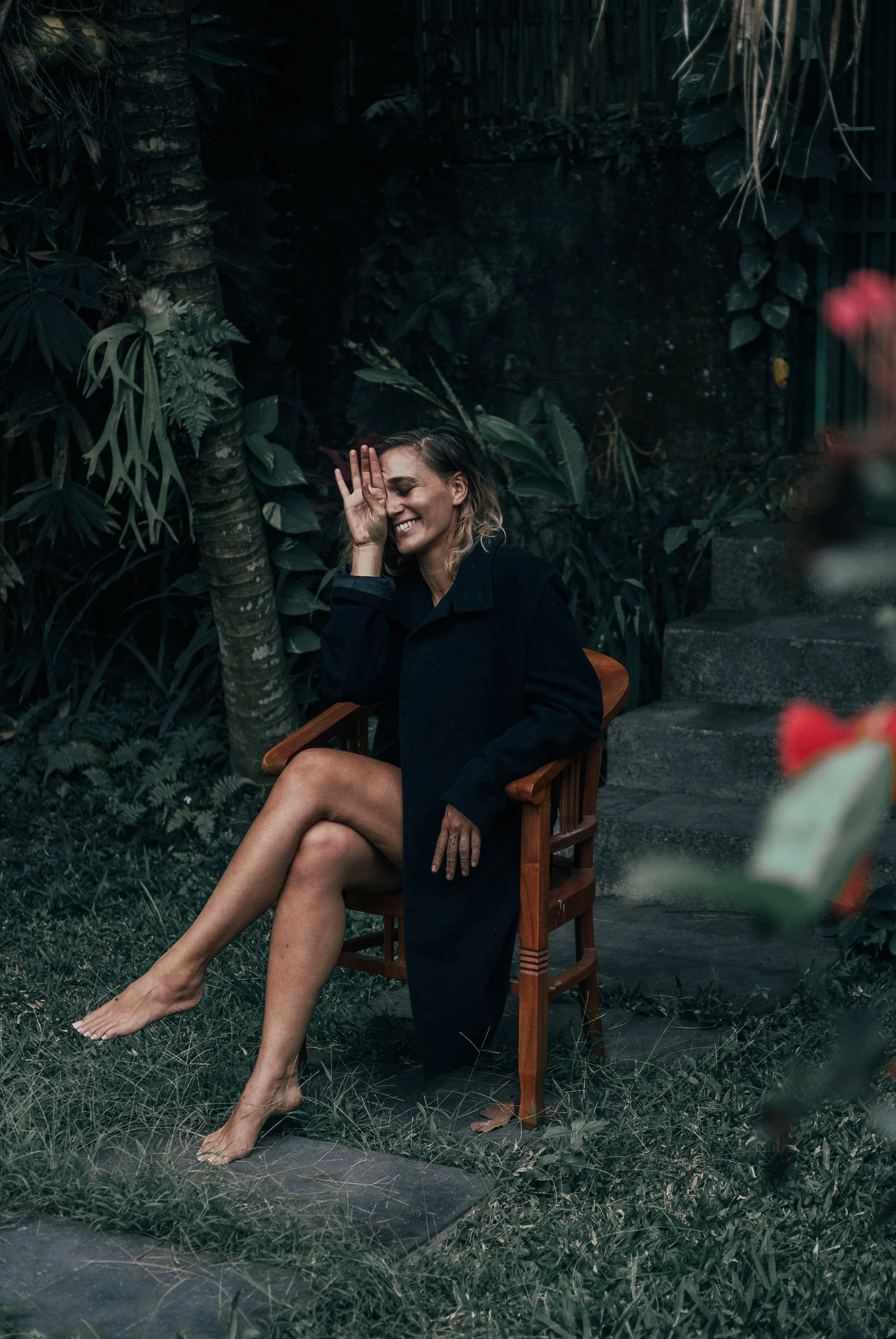 A smiling woman in a dark coat sits barefoot on a wooden chair in a lush green garden, covering part of her face with her hand.