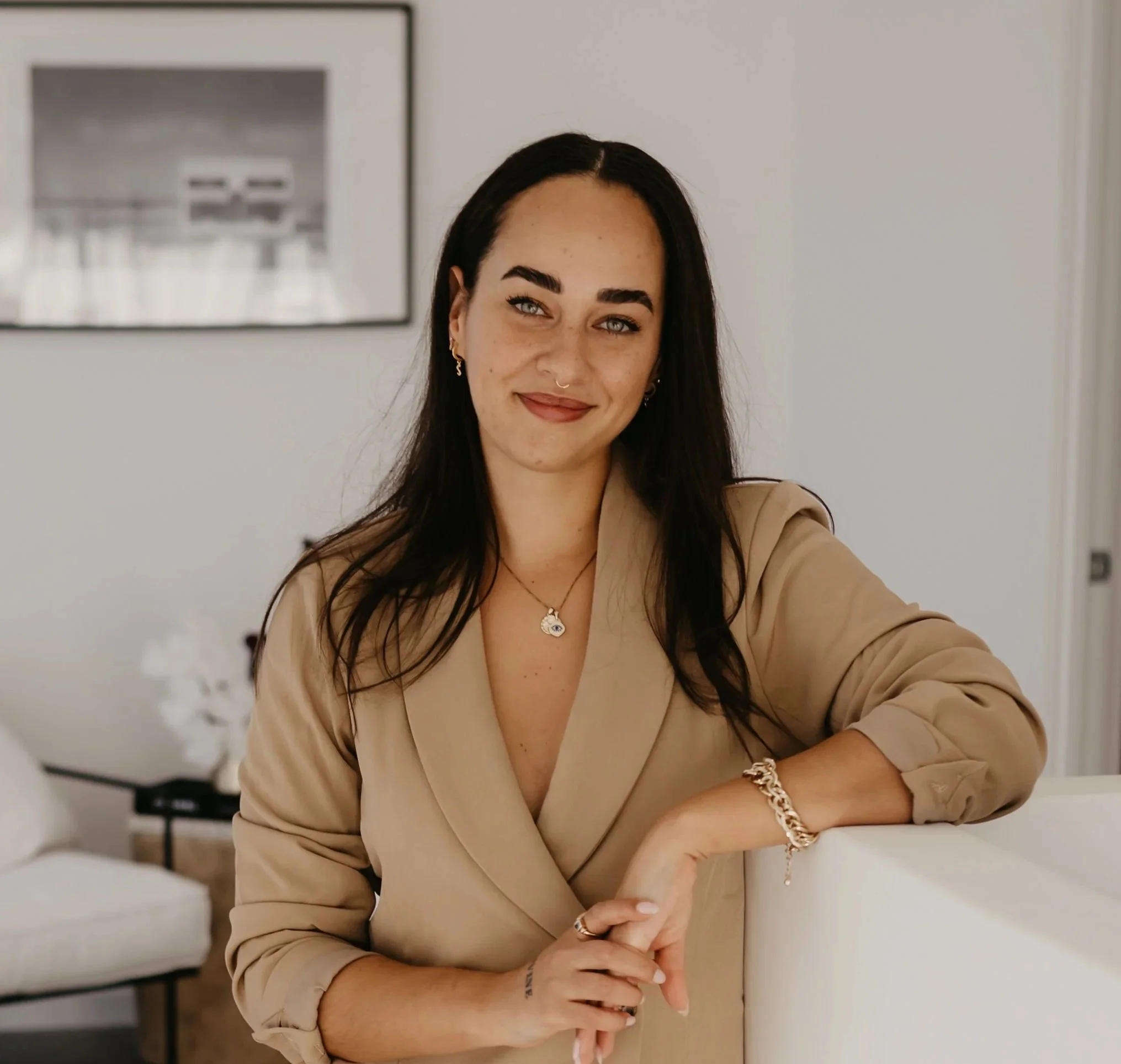 A woman with dark hair, light blue eyes, and light skin leaning on a white surface inside a modern, softly lit room. She is smiling and wearing a beige blazer, gold jewelry, and makeup.