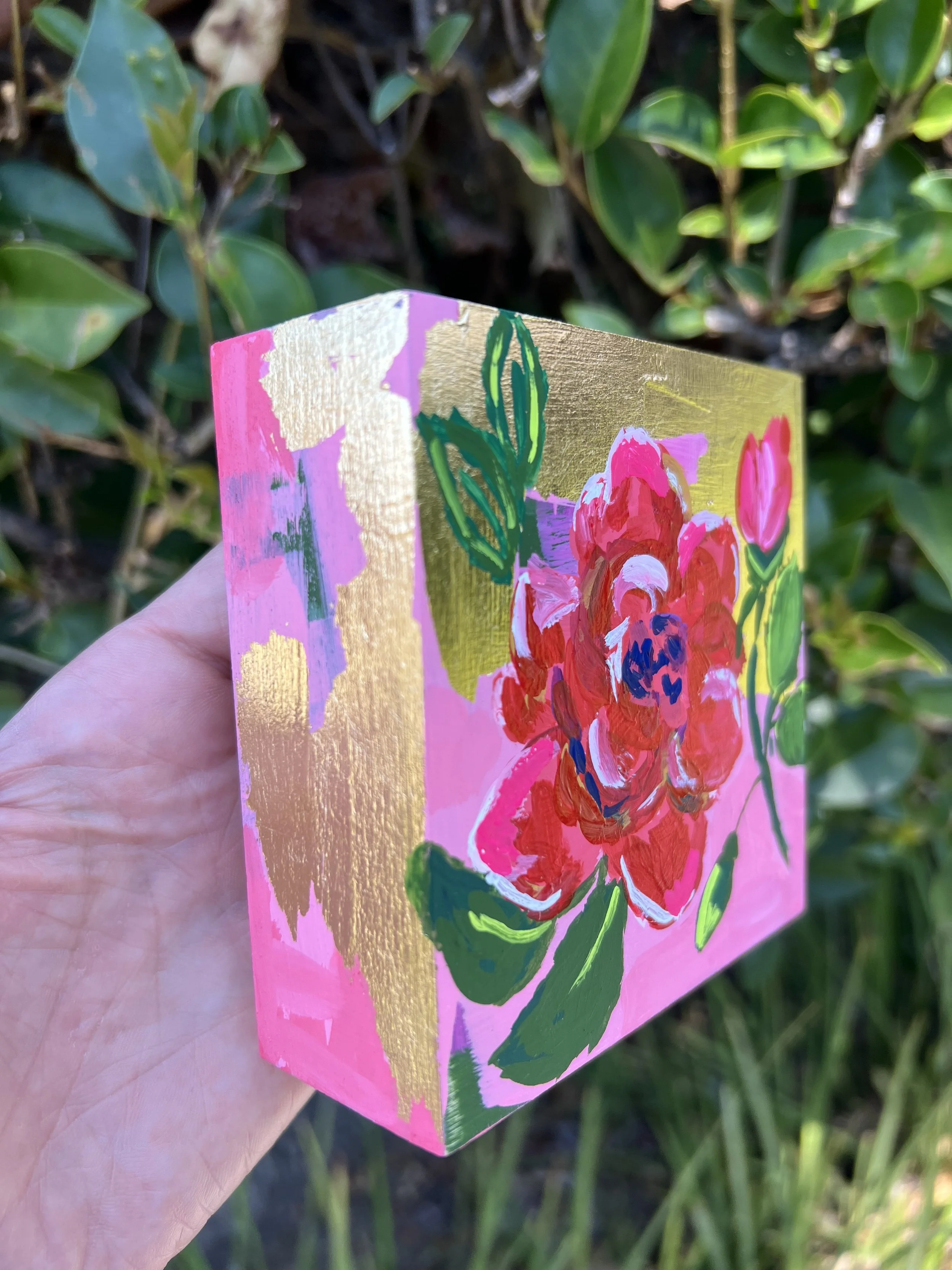 Close-up acrylic painting of a large red-pink rose flower and bud with dark blue center, accompanied by green foliage against gold leafing and pink abstract background.