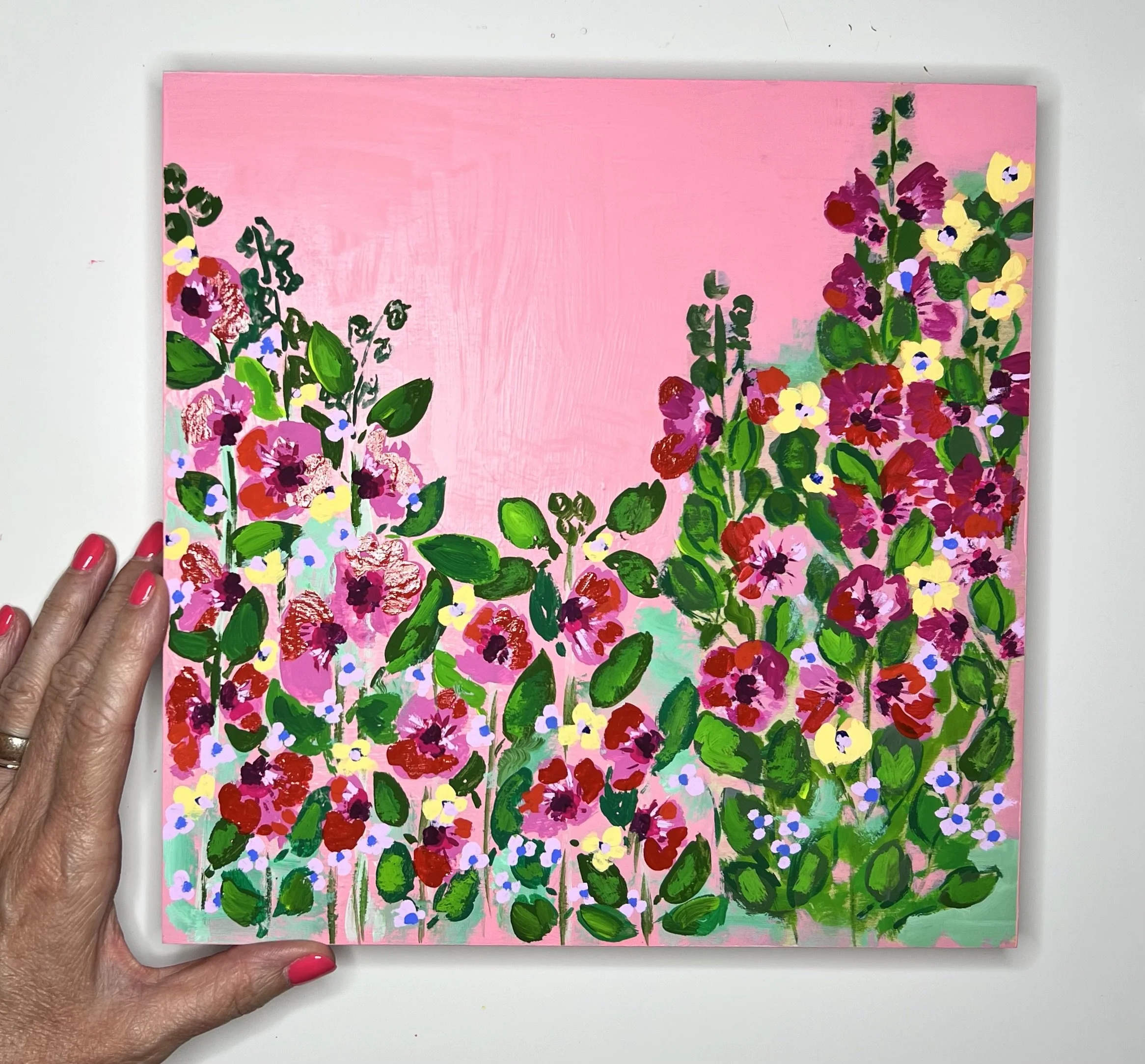 Vibrant hollyhocks painting featuring red, pink, and yellow blooms with lush green foliage against a soft pink background, capturing the joyful essence of an English spring garden in full bloom.