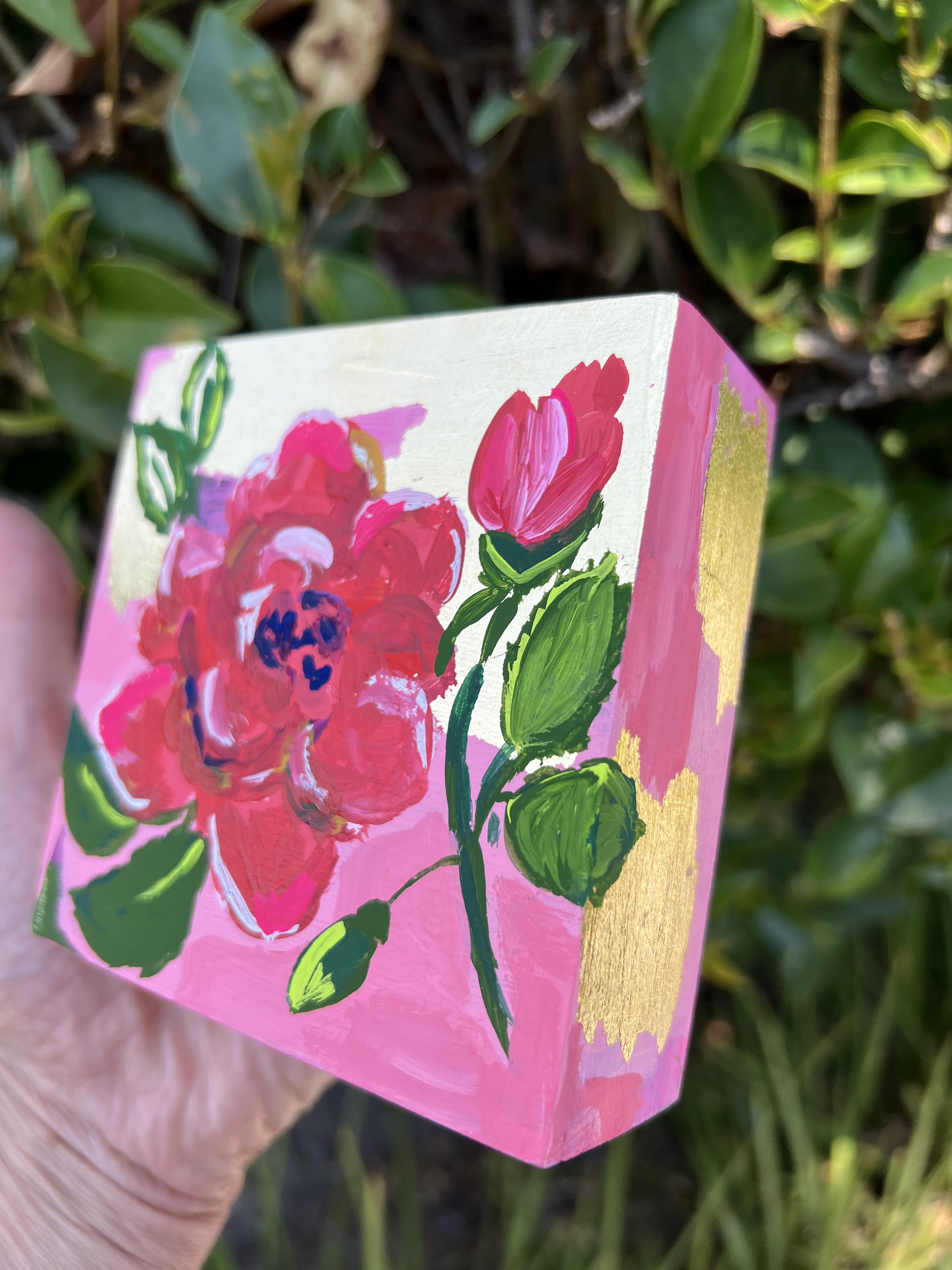 Close-up acrylic painting of a large red-pink rose flower and bud with dark blue center, accompanied by green foliage against gold leafing and pink abstract background.