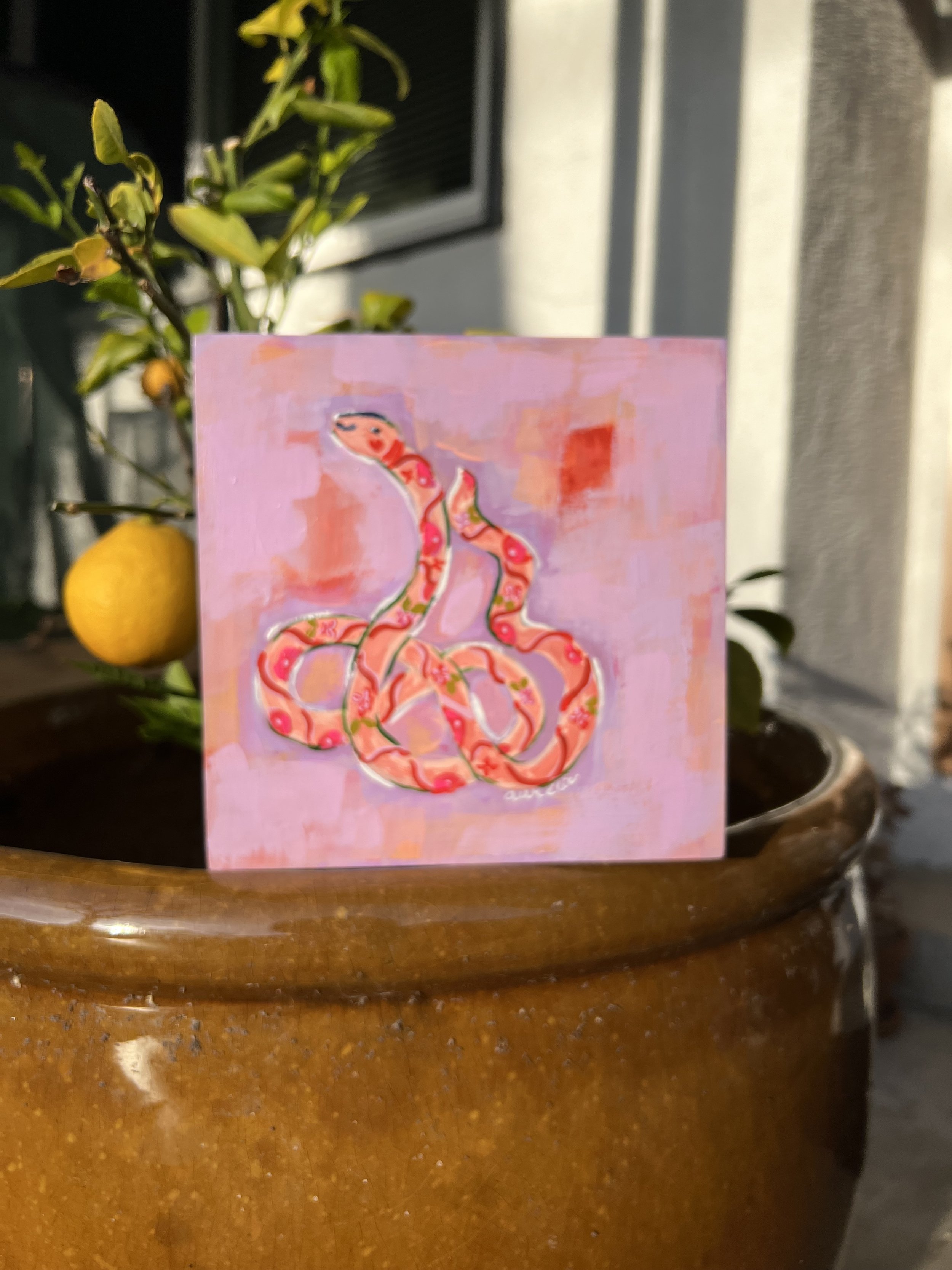 Playful, whimsical, flower-adorned snake that curves against a abstract lavender background. Delicate floral patterns and outlined in crisp white lines, approachable and feminine