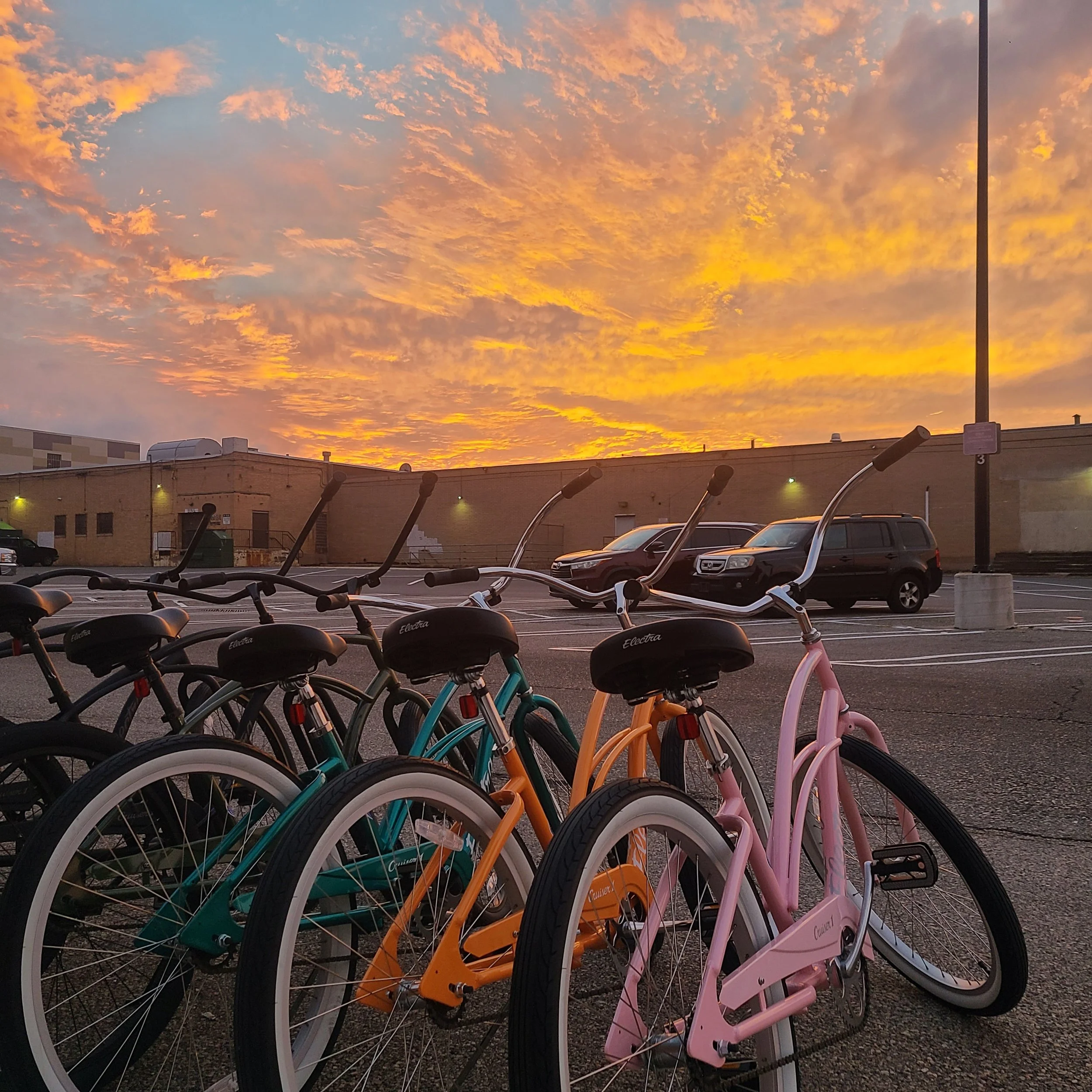 A row of colorful Electra beach cruiser bicycles parked in a parking lot silhouetted against a brilliant orange and yellow sunset.