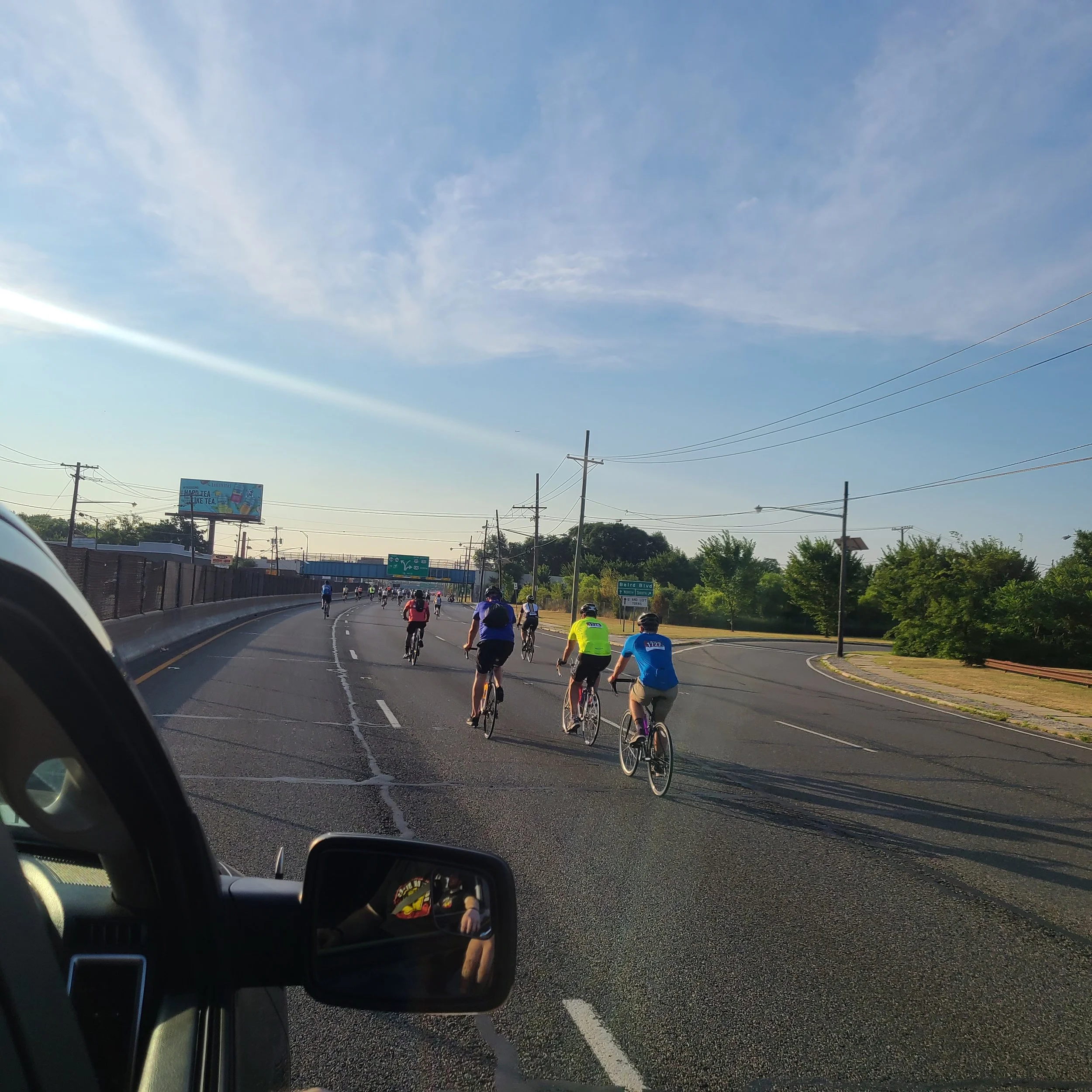 Cyclists pedaling down a wide highway road as captured from a support vehicle window during a local charity ride in Philadelphia.