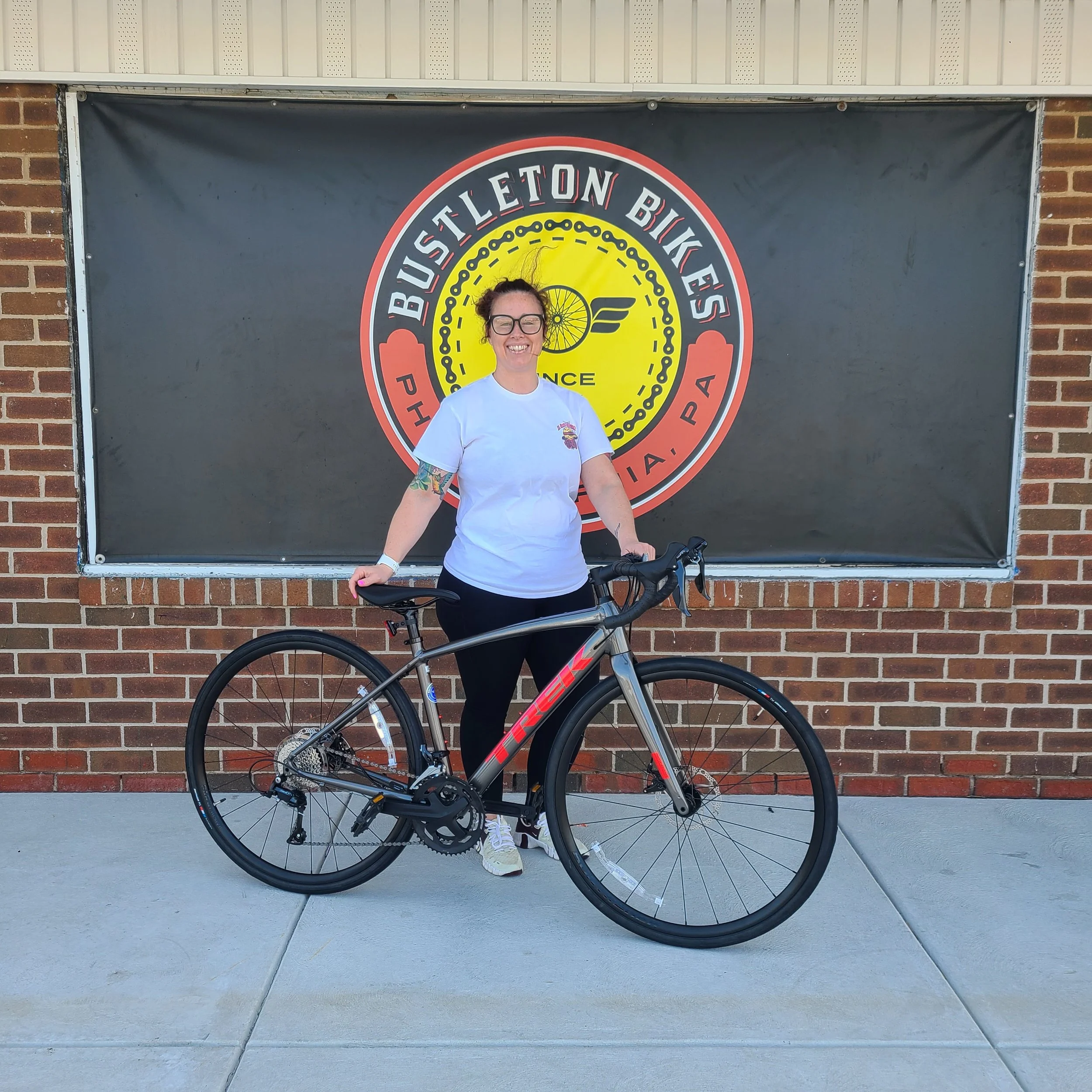 A smiling woman with tattoos standing outside with a dark grey Trek Domane aluminum endurance road bike with red logos.