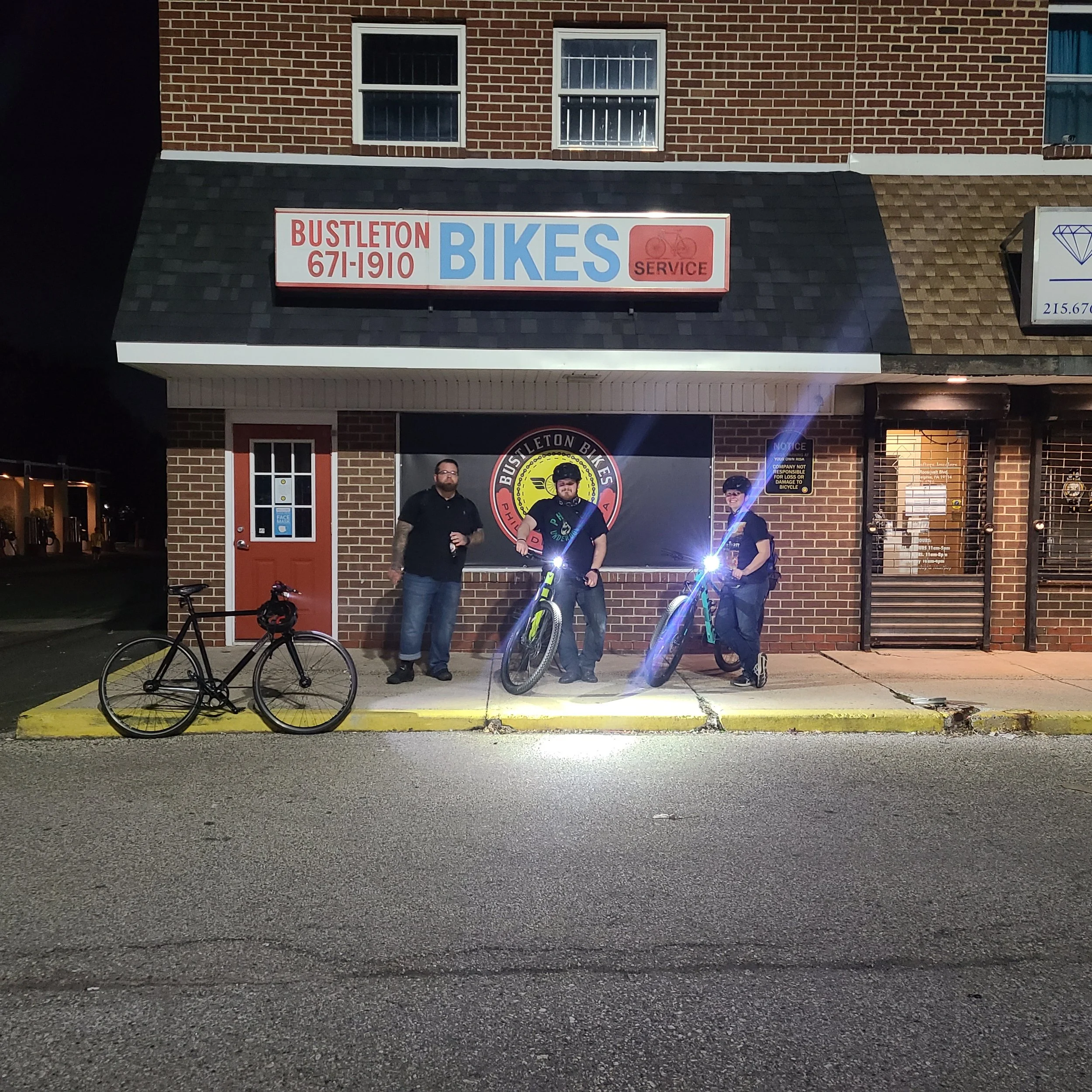 Three cyclists with high-powered bike headlights illuminated standing outside the storefront at night before a group ride.