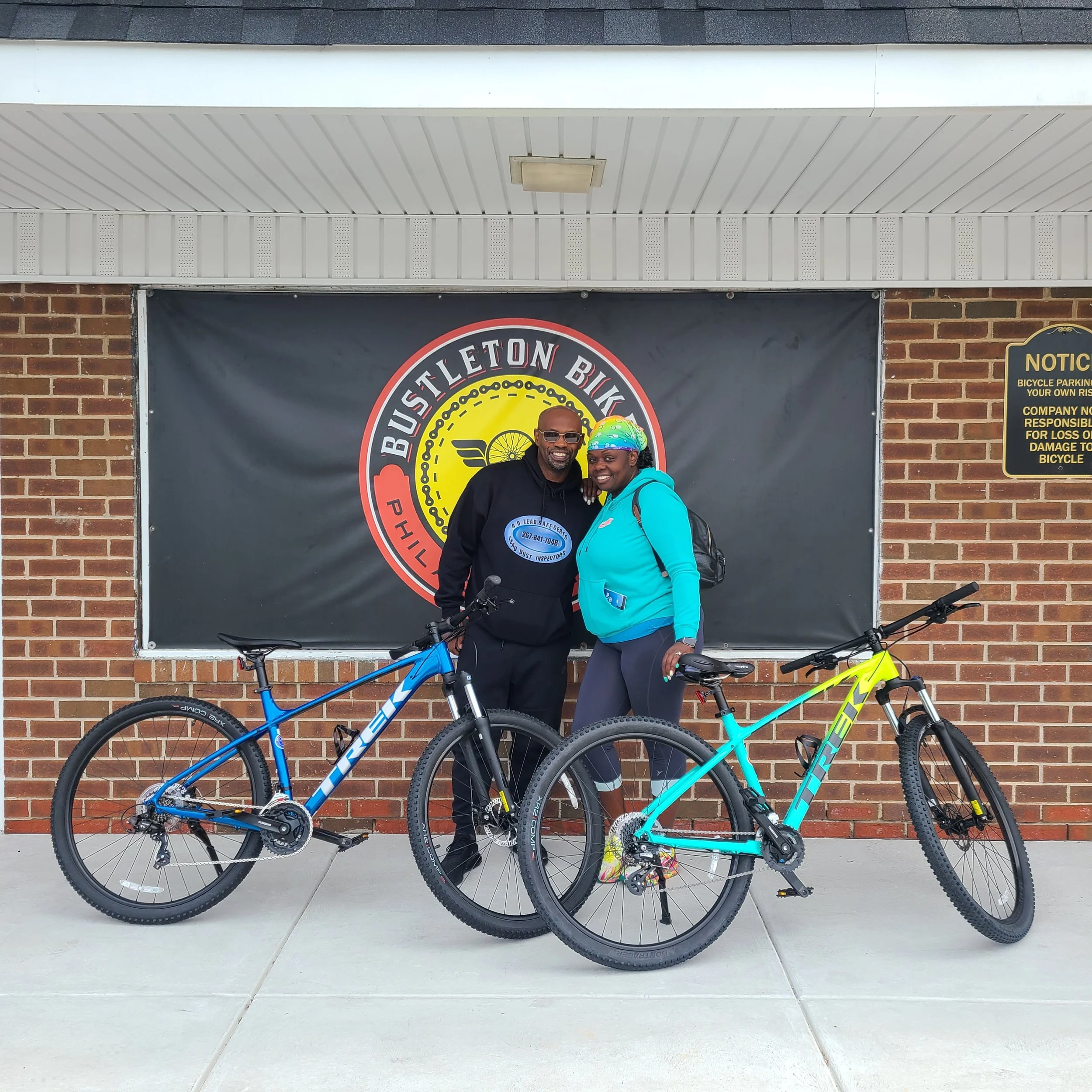 A happy couple smiling and posing together with their two newly purchased Trek hybrid bicycles in front of the shop's banner.