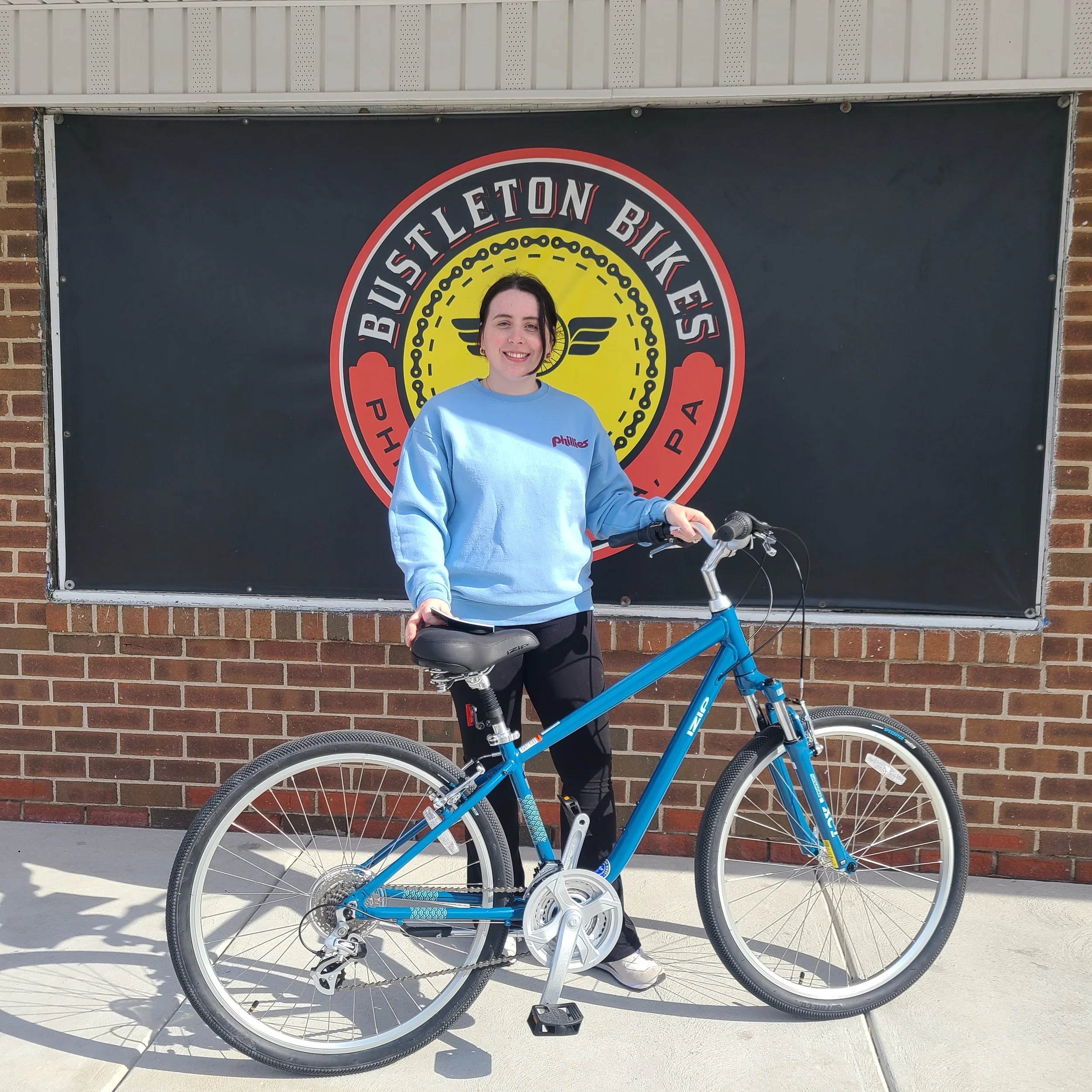 A smiling young woman wearing a blue Phillies sweatshirt posing outside with a bright blue step-through comfort hybrid bicycle.