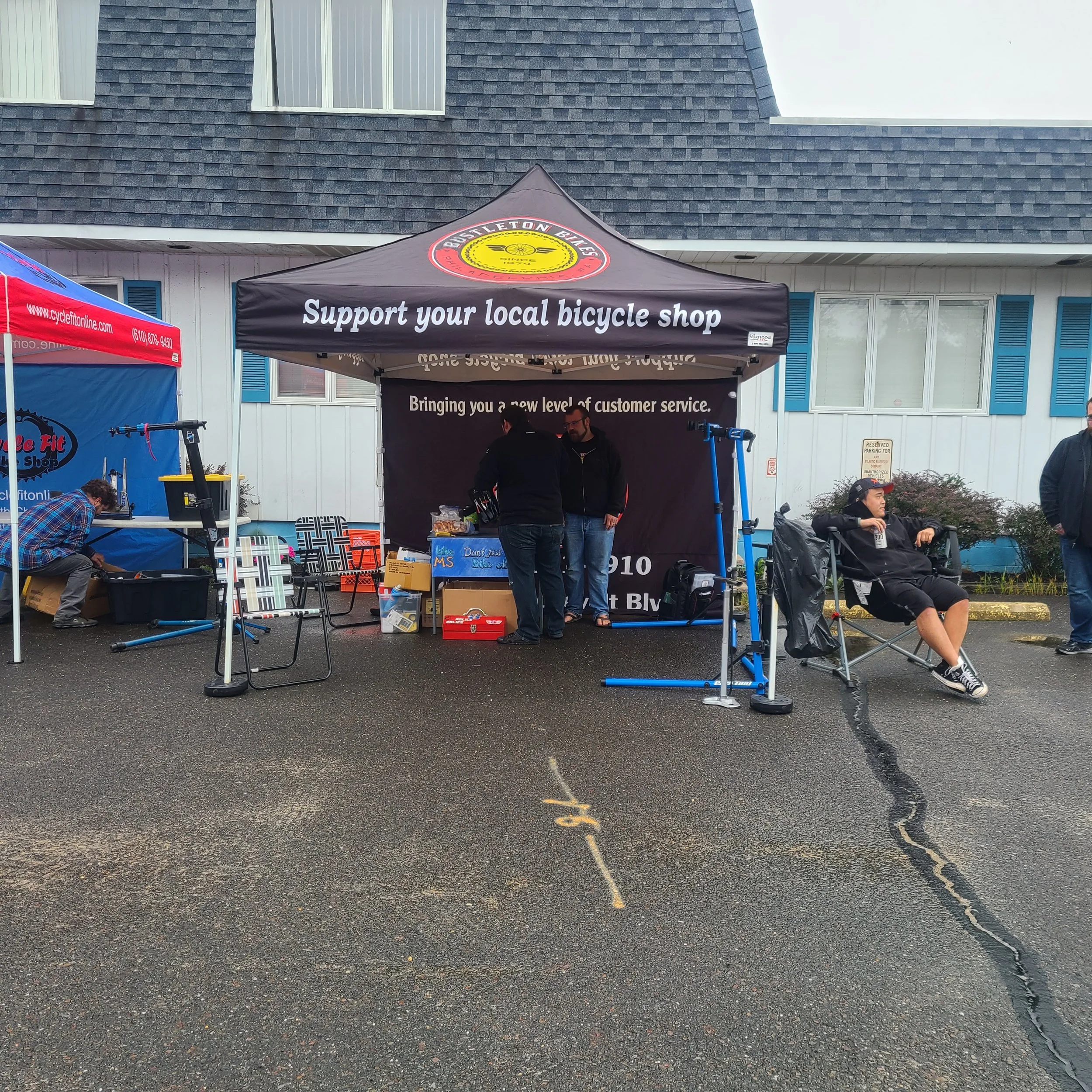 The black Bustleton Bikes canopy tent setup in a parking lot, reading 'Support your local bicycle shop' with repair stands ready for action