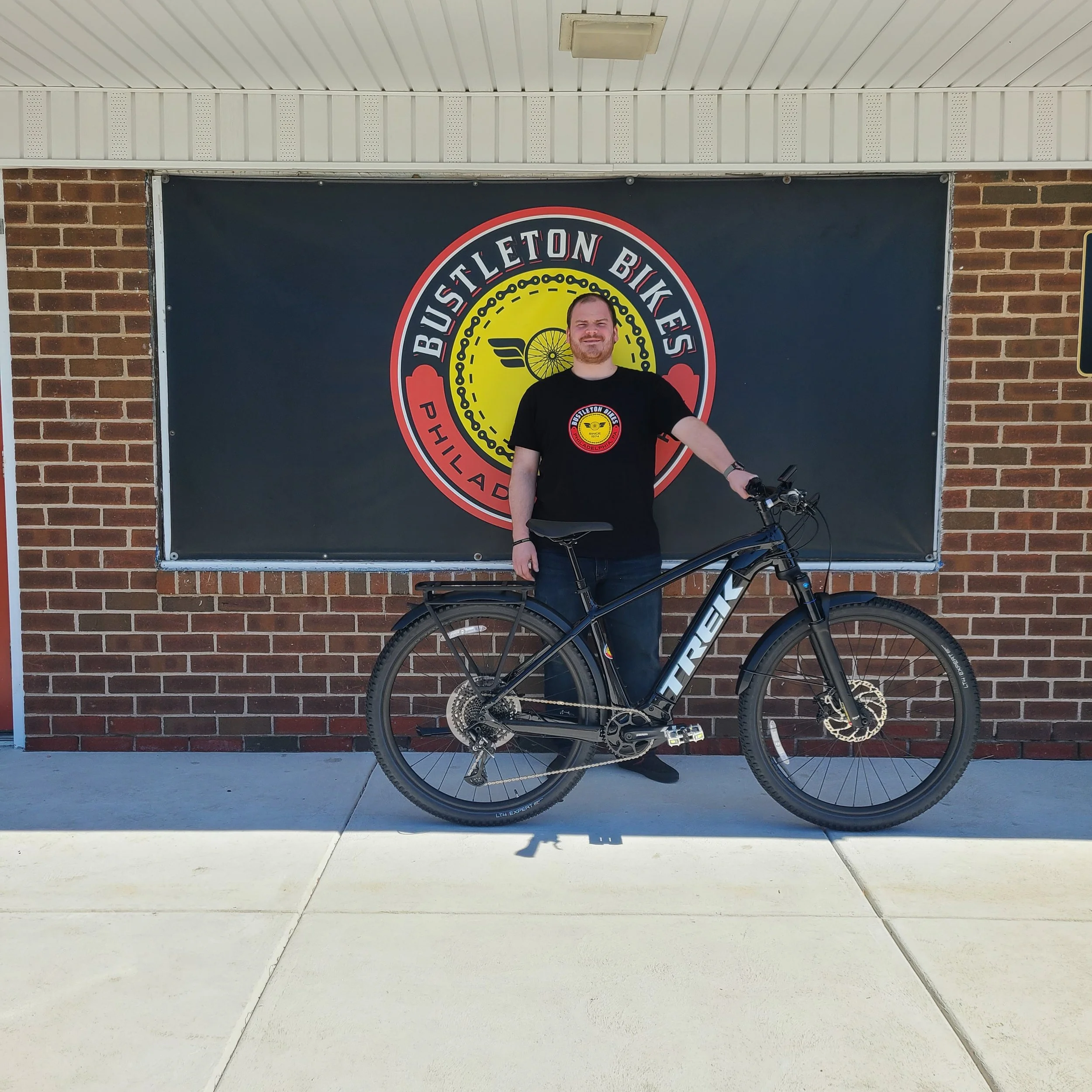 Jordan smiling while standing proudly with his new black Trek Powerfly electric mountain bike equipped with fenders and a rear cargo rack.