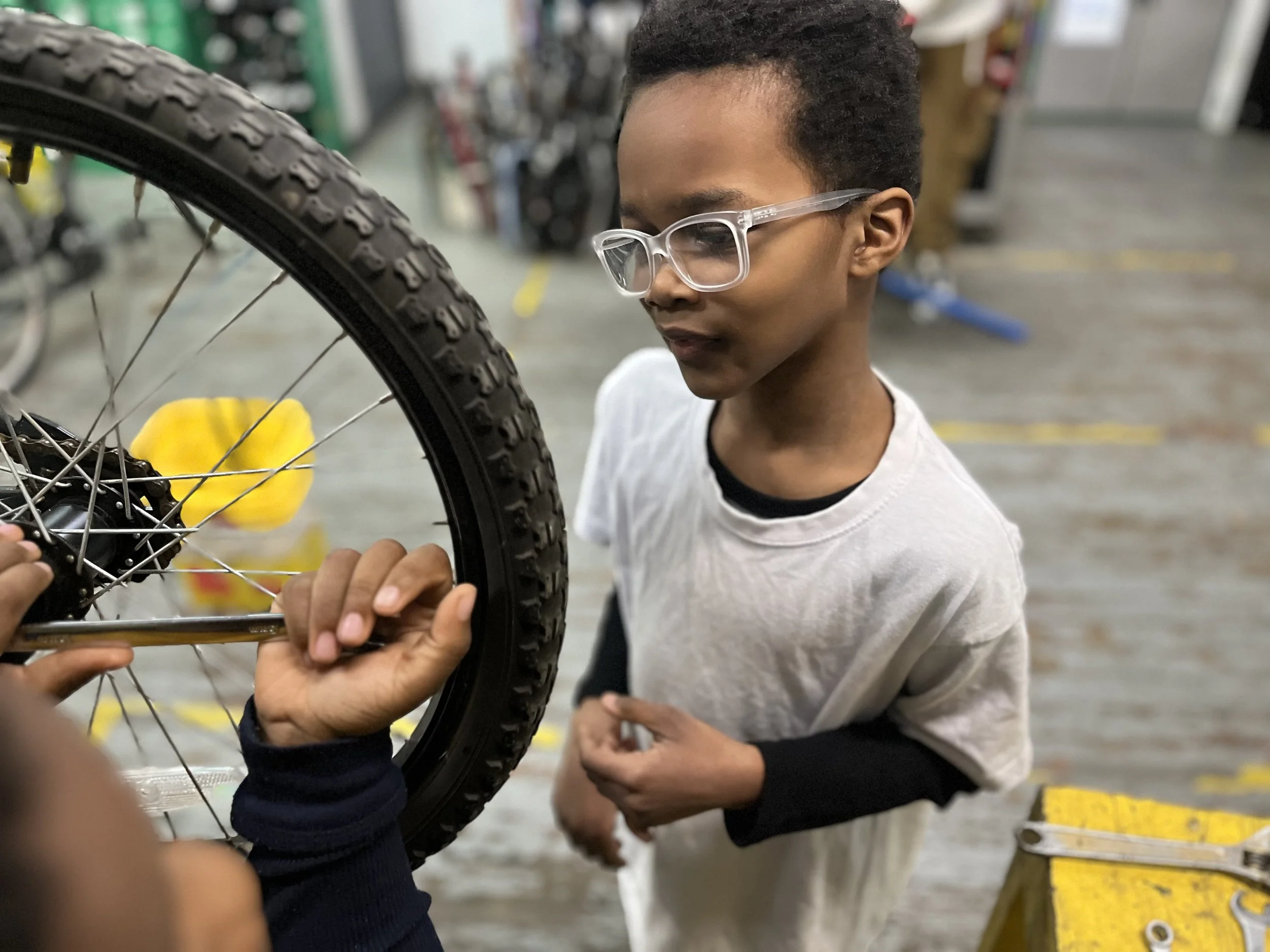 A young boy with glasses closely observing a bicycle wheel being repaired at Neighborhood Bike Works in Philadelphia.