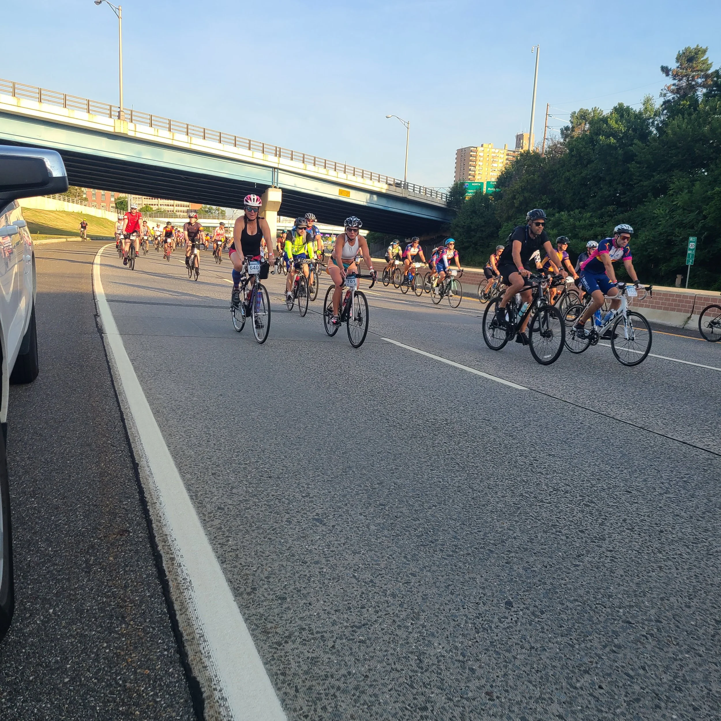 A large pack of cyclists wearing helmets and jerseys riding down an open highway during the Ben to the Shore Bike Tour under a clear morning sky