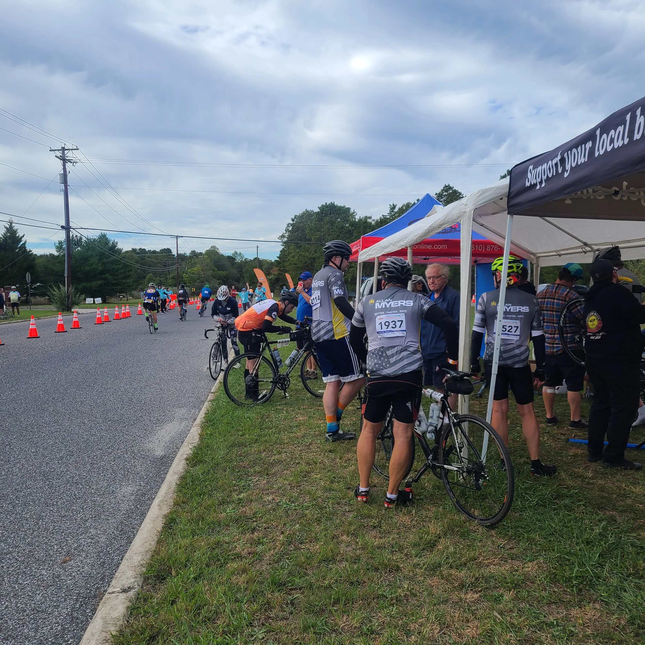Cyclists wearing jerseys standing near a roadside team tent with their bikes during the MS City to Shore charity ride