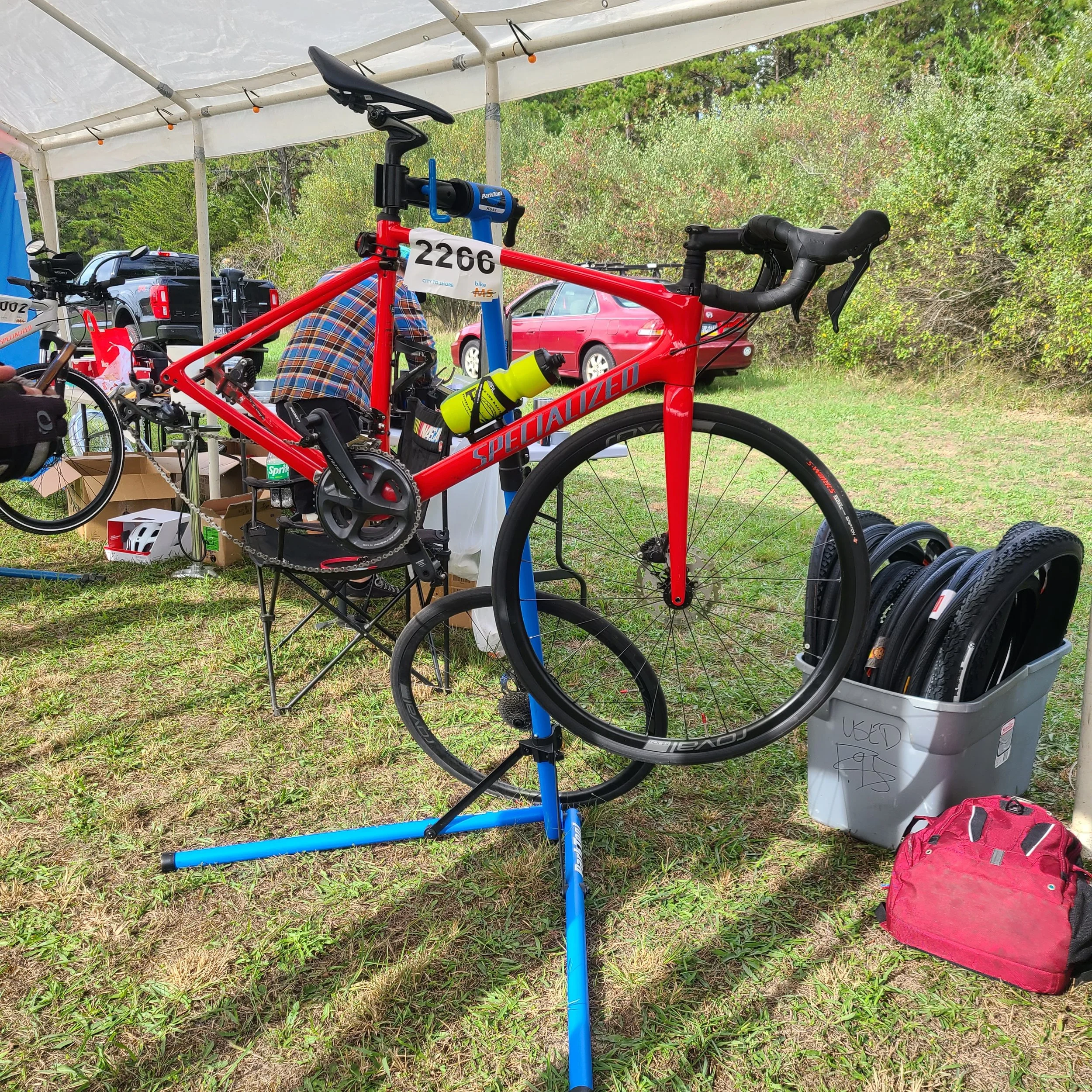 A bright red Specialized road bike mounted on a blue Park Tool repair stand under a tent at an MS City to Shore rest stop.