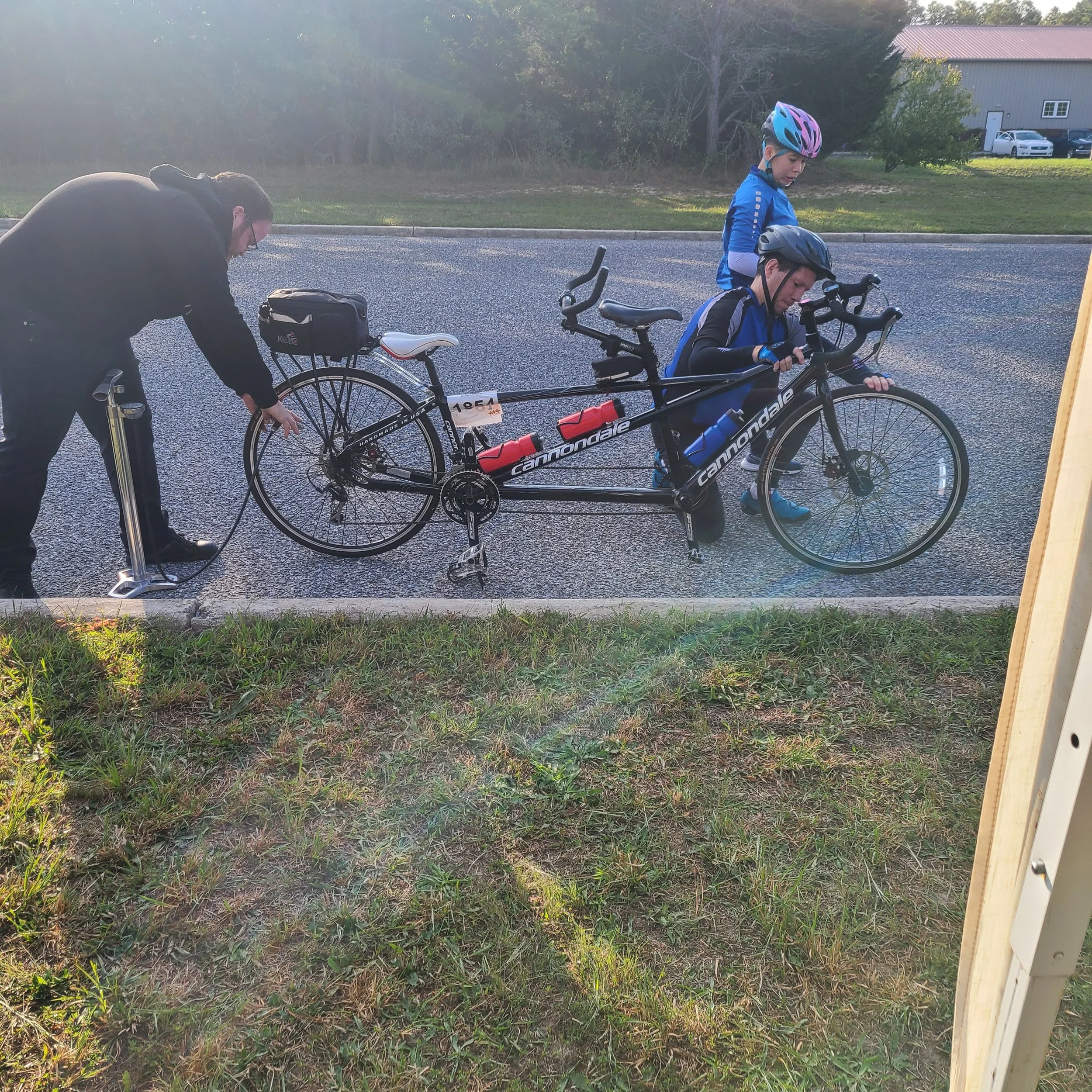 A mechanic uses a floor pump to inflate the rear tire of a long black Cannondale tandem bicycle during a charity cycling event.