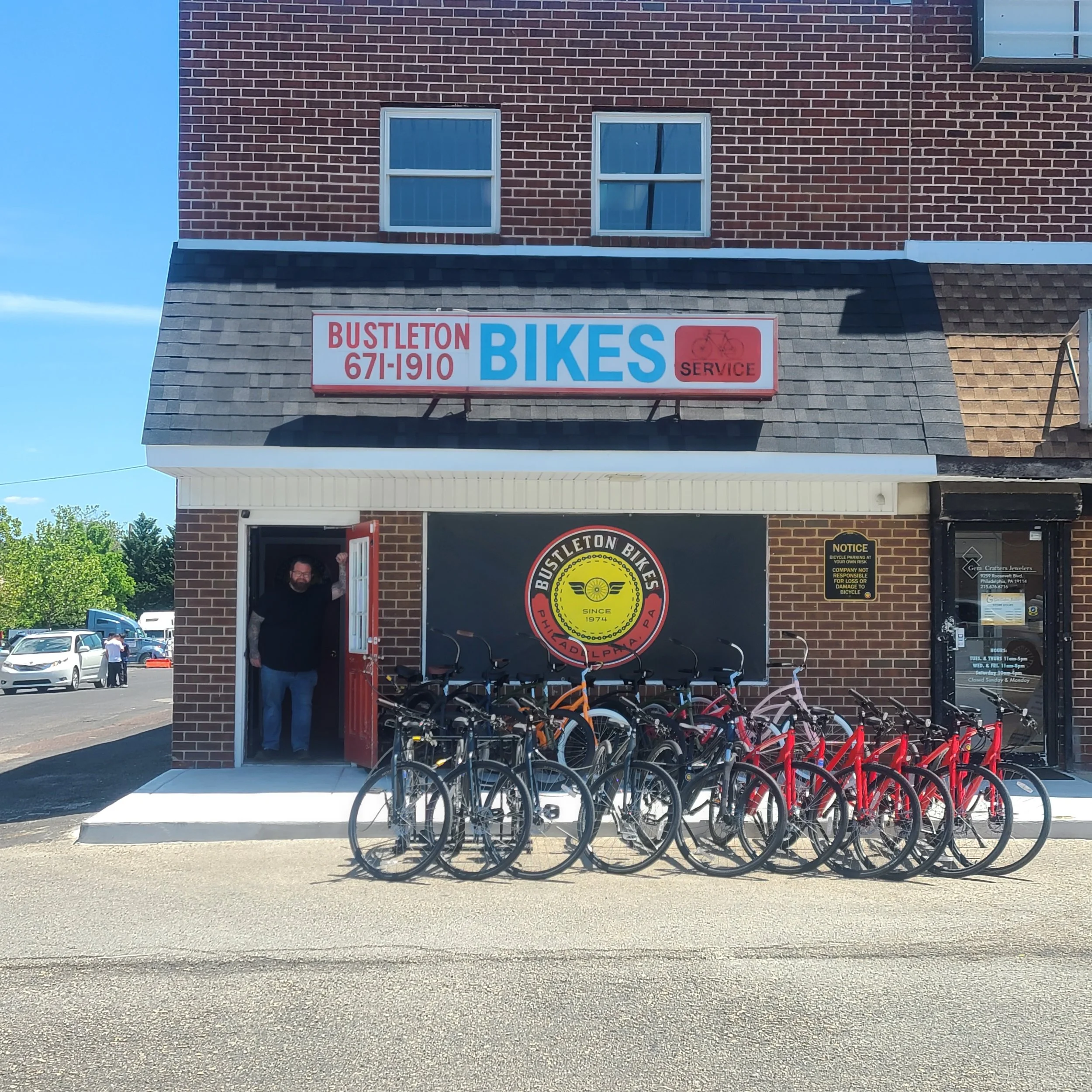 The owner of Bustleton Bikes stands welcomingly in the doorway of the brick storefront in Philadelphia, with a row of bicycles parked out front.