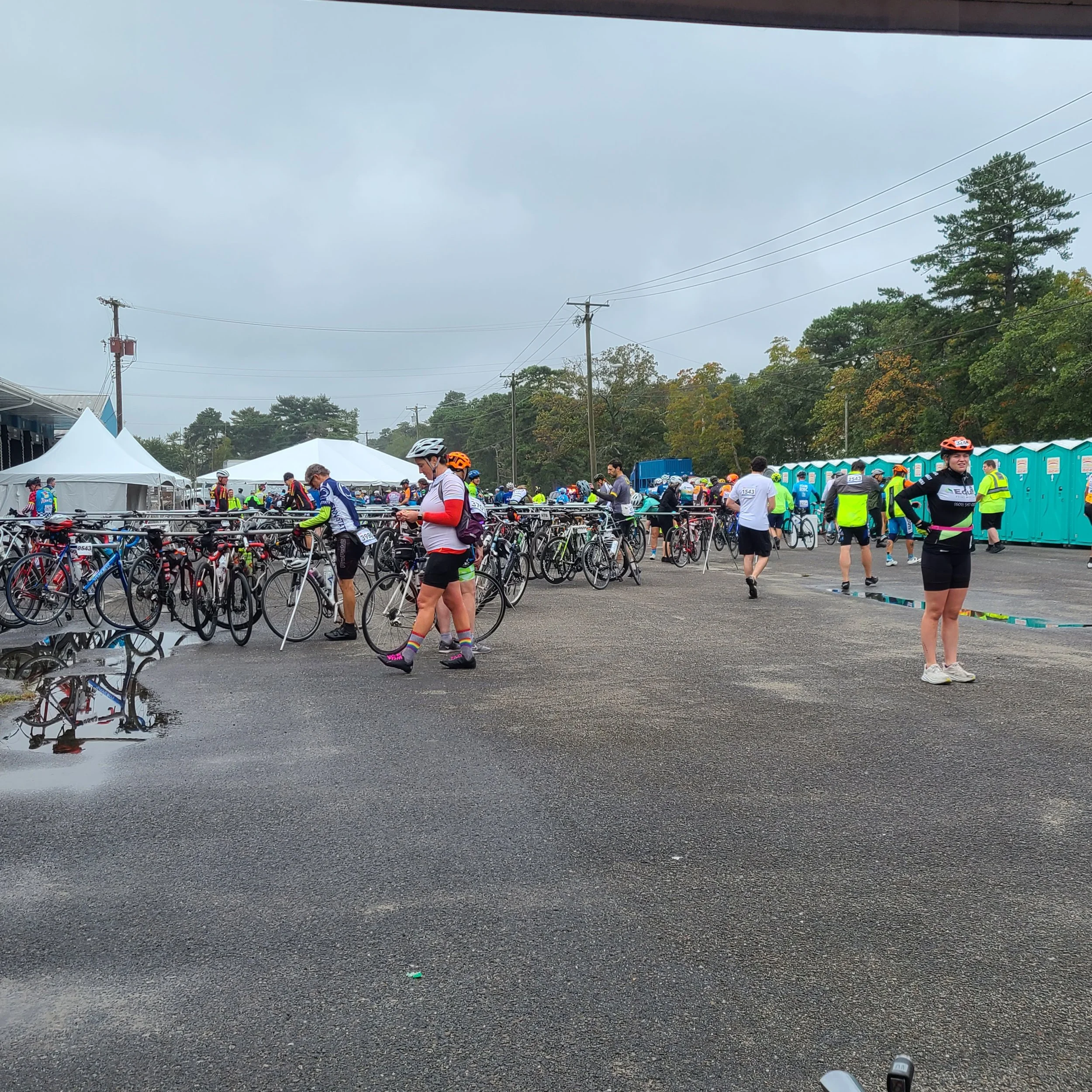 A busy outdoor rest stop area filled with bicycle racks, resting cyclists, and event tents on an overcast day during the MS City to Shore charity ride.