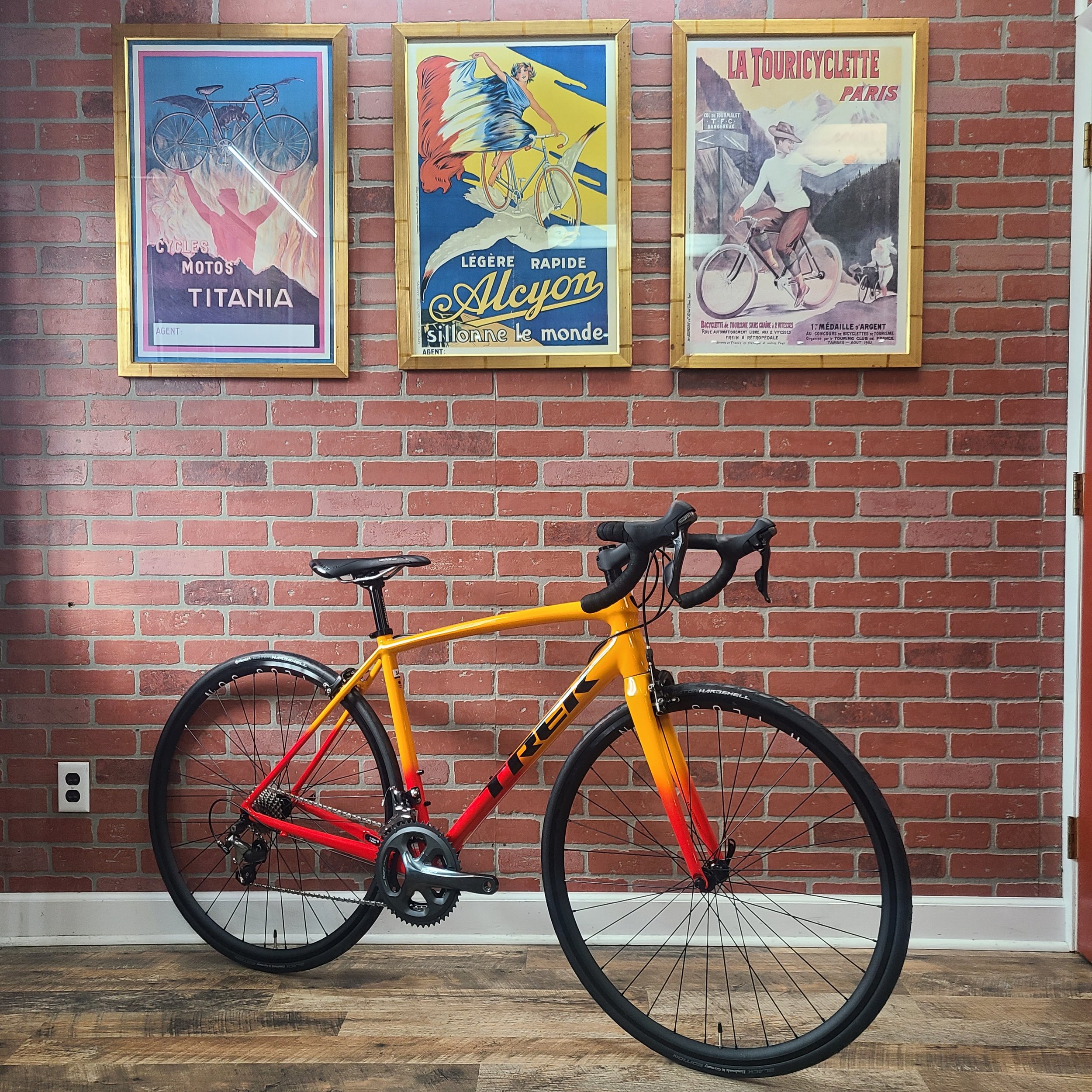 A custom-built Trek Émonda ALR 5 road bike with a vibrant yellow-to-red fade frame parked in front of a brick wall decorated with vintage cycling posters.