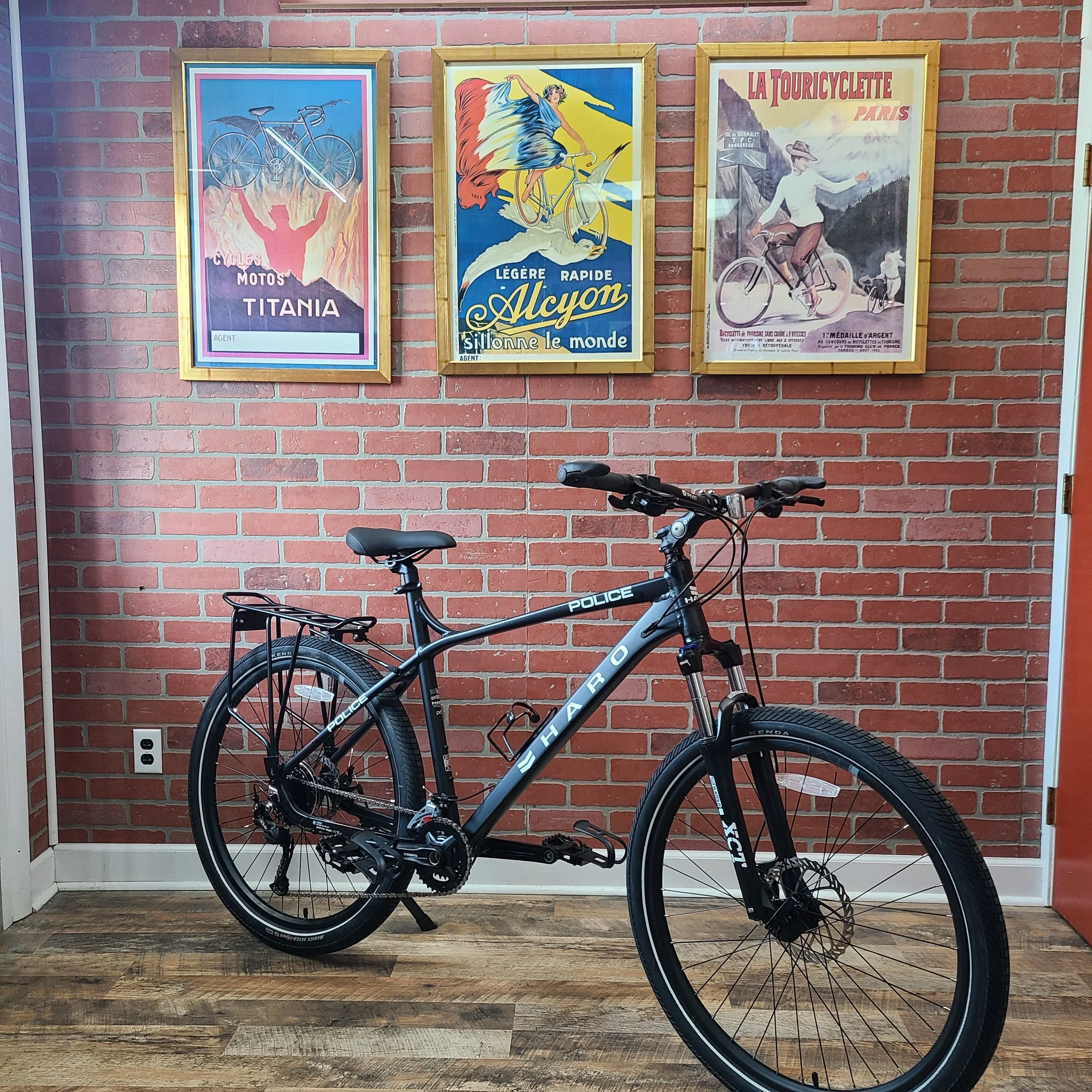 A matte black Haro Police edition mountain bike with a rear rack displayed against a brick wall beneath vintage French cycling posters.