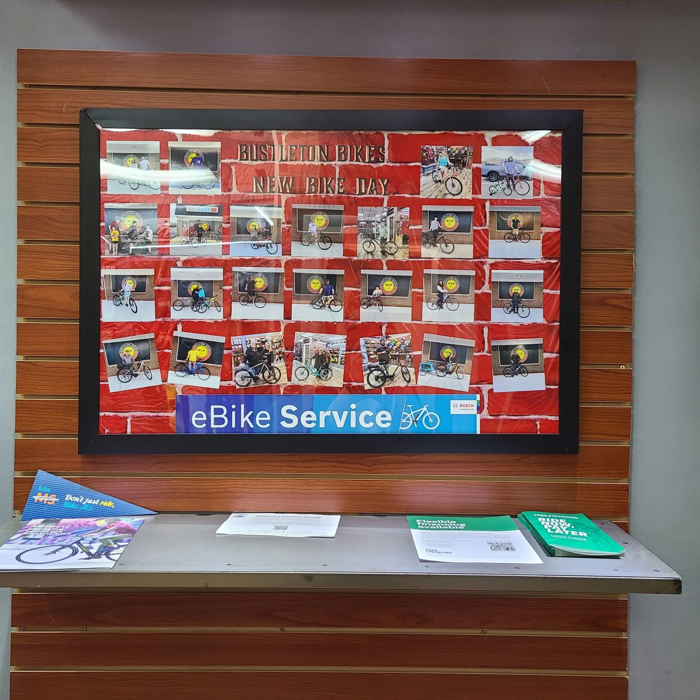 A framed collage titled "Bustleton Bikes New Bike Day" hanging on a wooden wall, filled with photos of smiling customers with their new bicycles.
