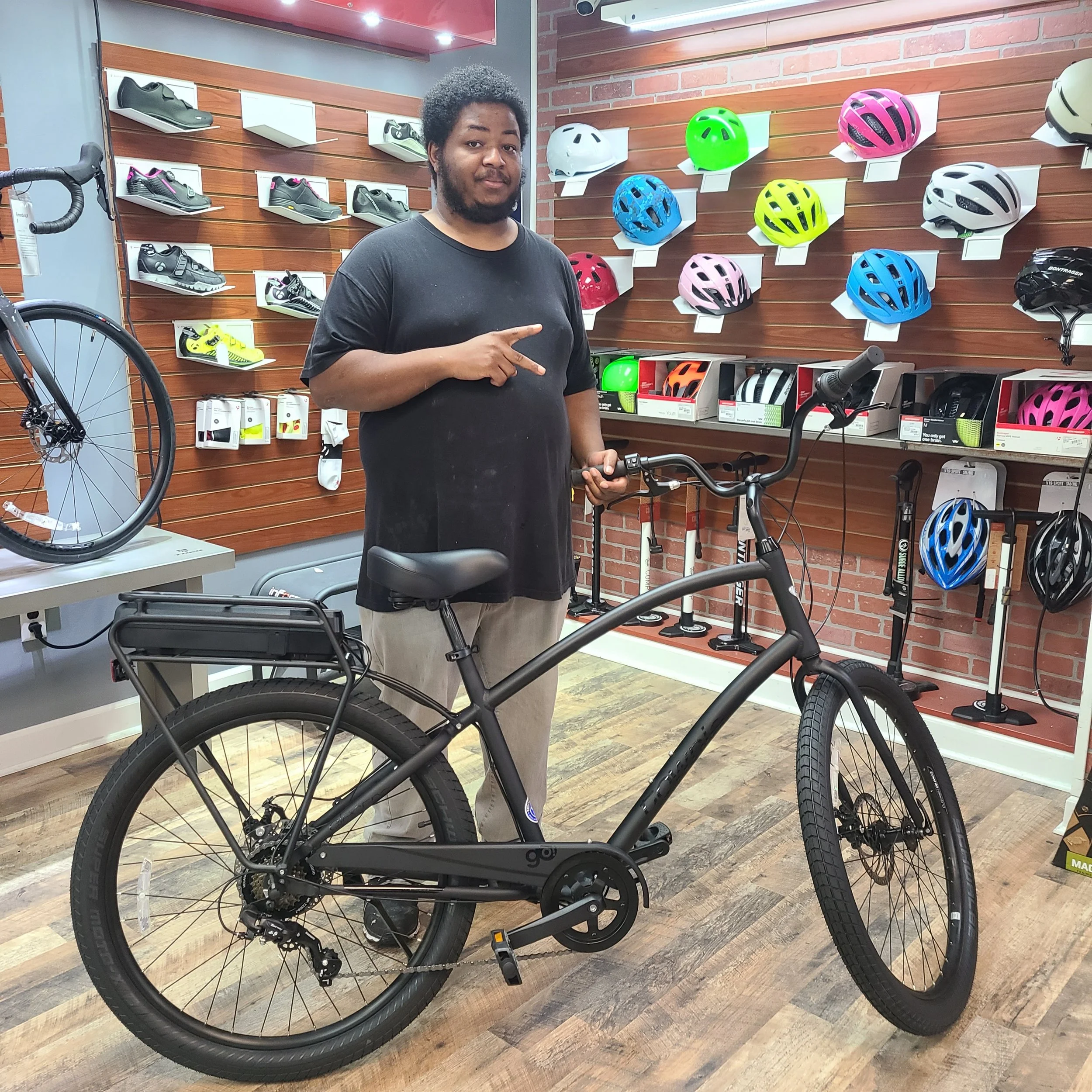 A young man throwing up a peace sign inside the bike shop display area next to a matte black Electra Townie Go cruiser e-bike.