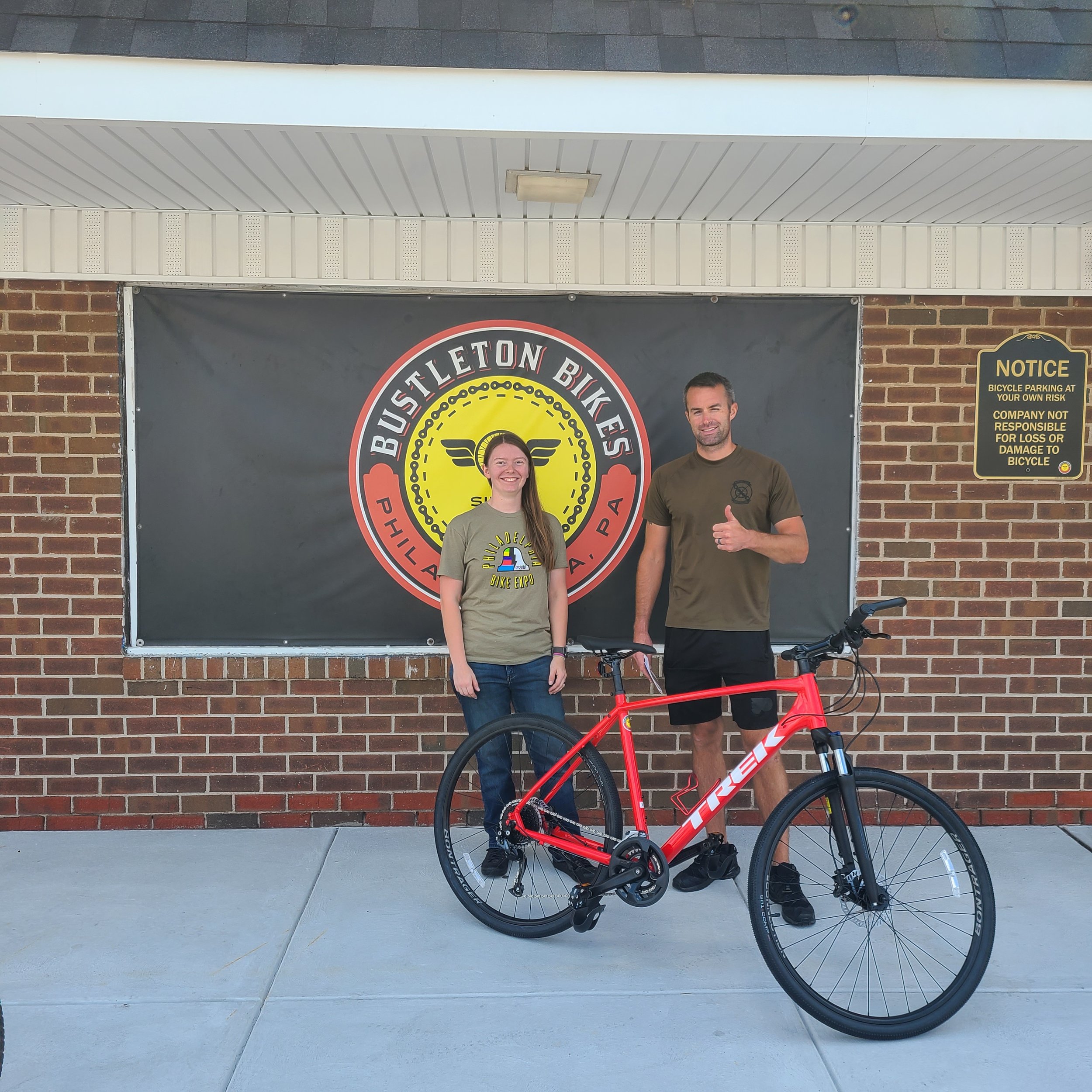 Me and a happy customer smile and pose together with a brand-new red Trek hybrid bike in front of the Bustleton Bikes storefront banner.
