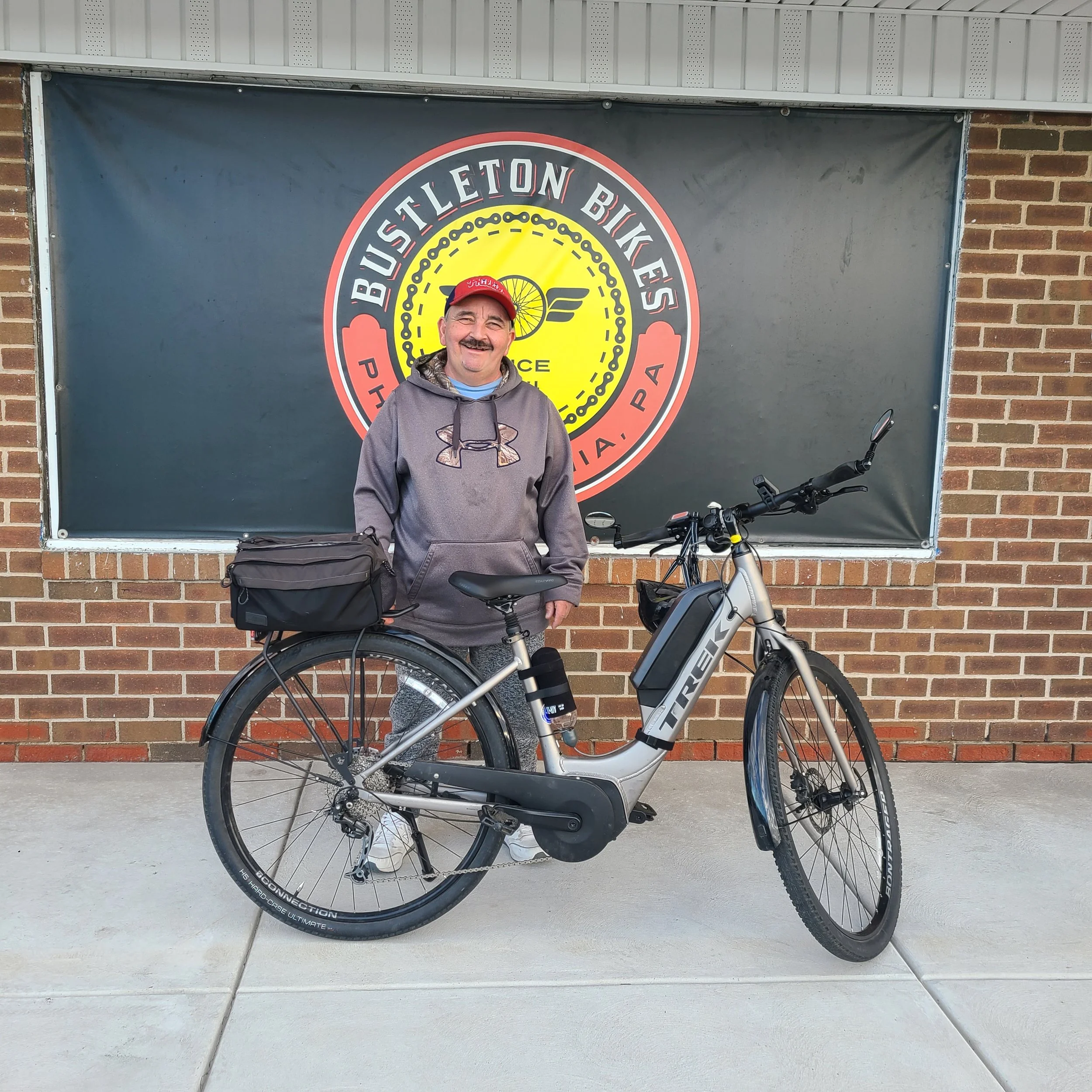 A man wearing a red cap standing proudly outside with a silver Trek Verve+ lowstep electric hybrid bicycle equipped with a rear commuter bag.