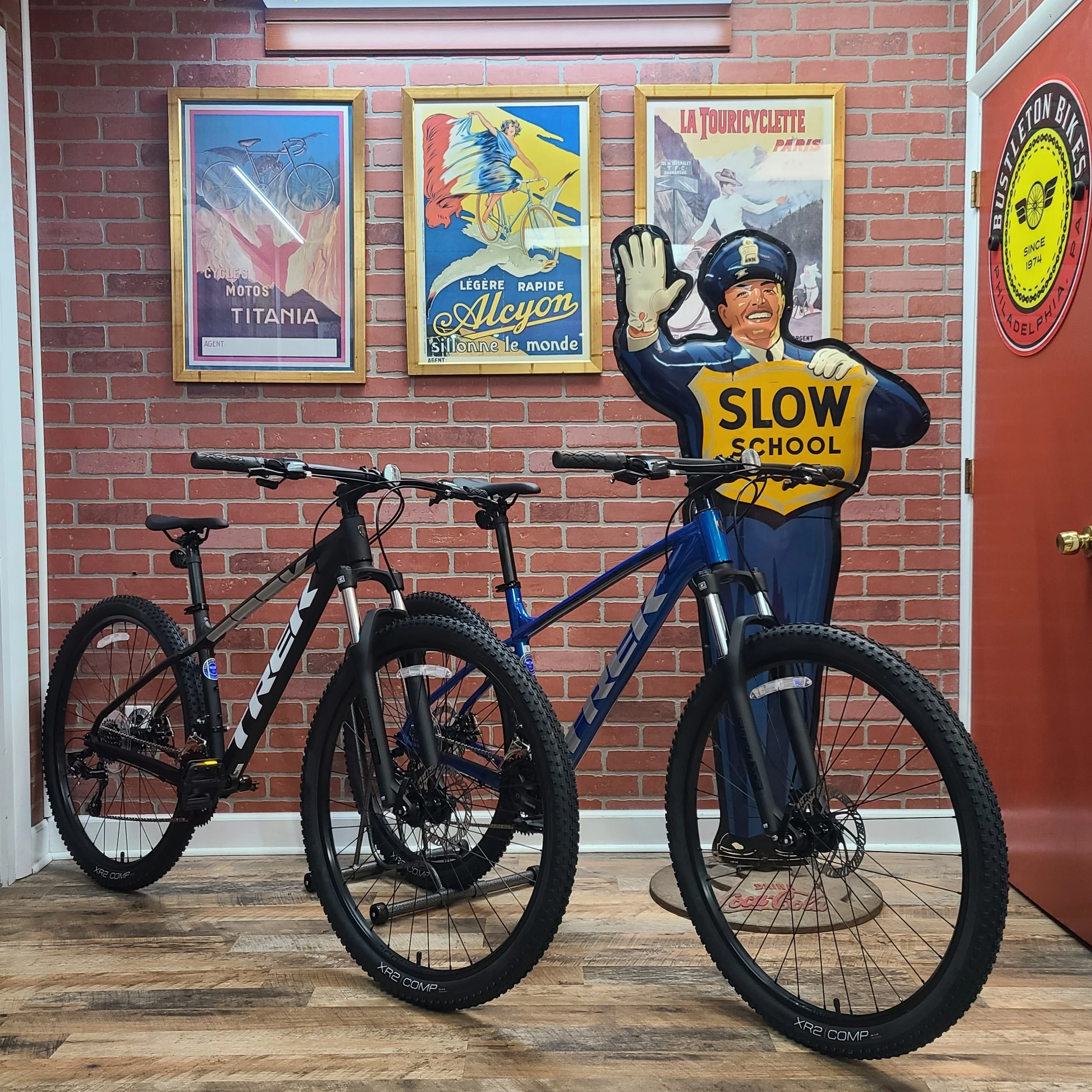 Two Trek Marlin 4 mountain bikes—one matte black and one royal blue—parked indoors in front of a vintage "Slow School" traffic cop sign on a brick wall.