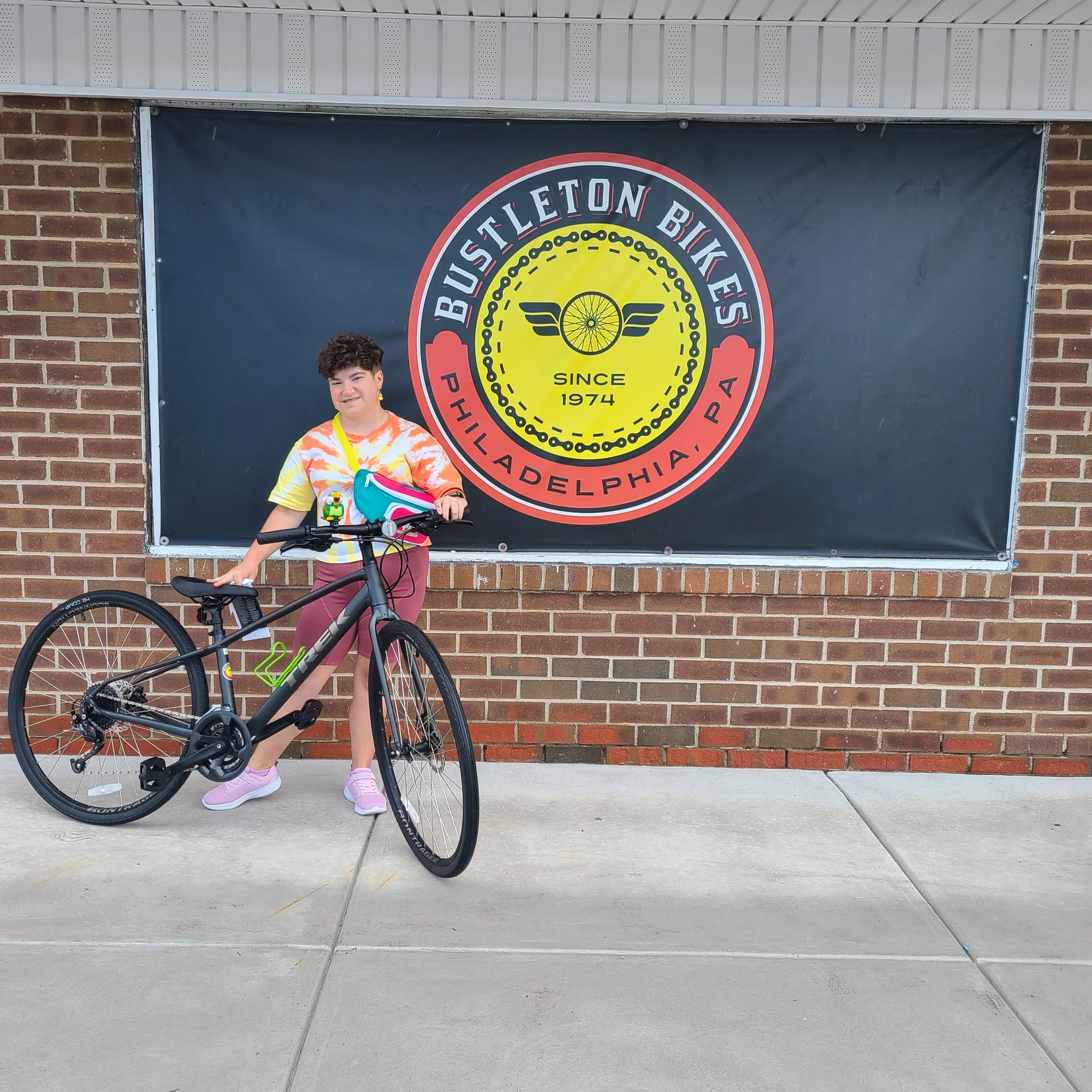 A young person in a tie-dye shirt standing outside the bike shop banner with a sleek matte black Trek FX hybrid bicycle.
