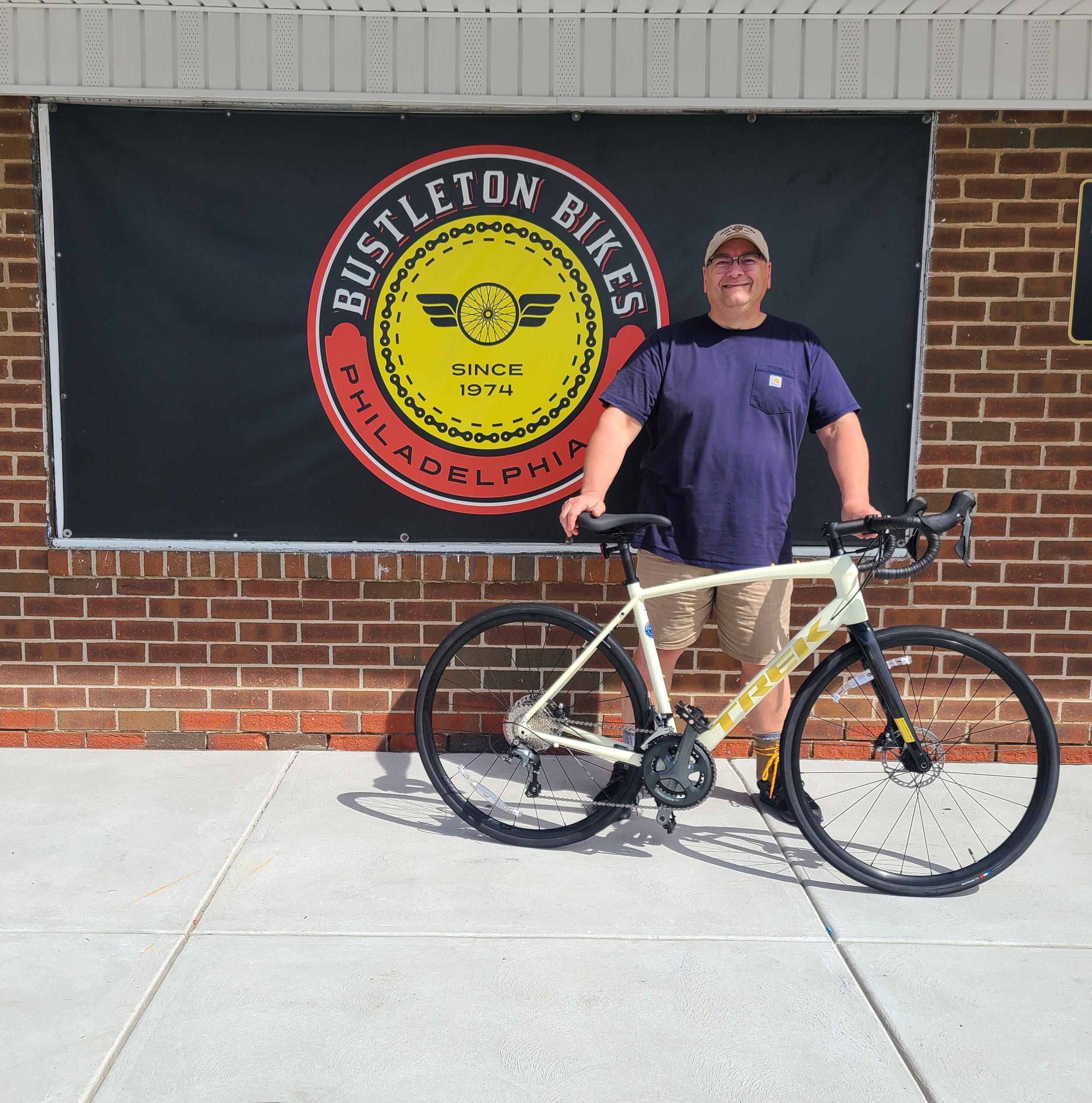 A smiling man in a navy t-shirt posing outside with a clean cream-colored Trek Domane AL endurance road bike.