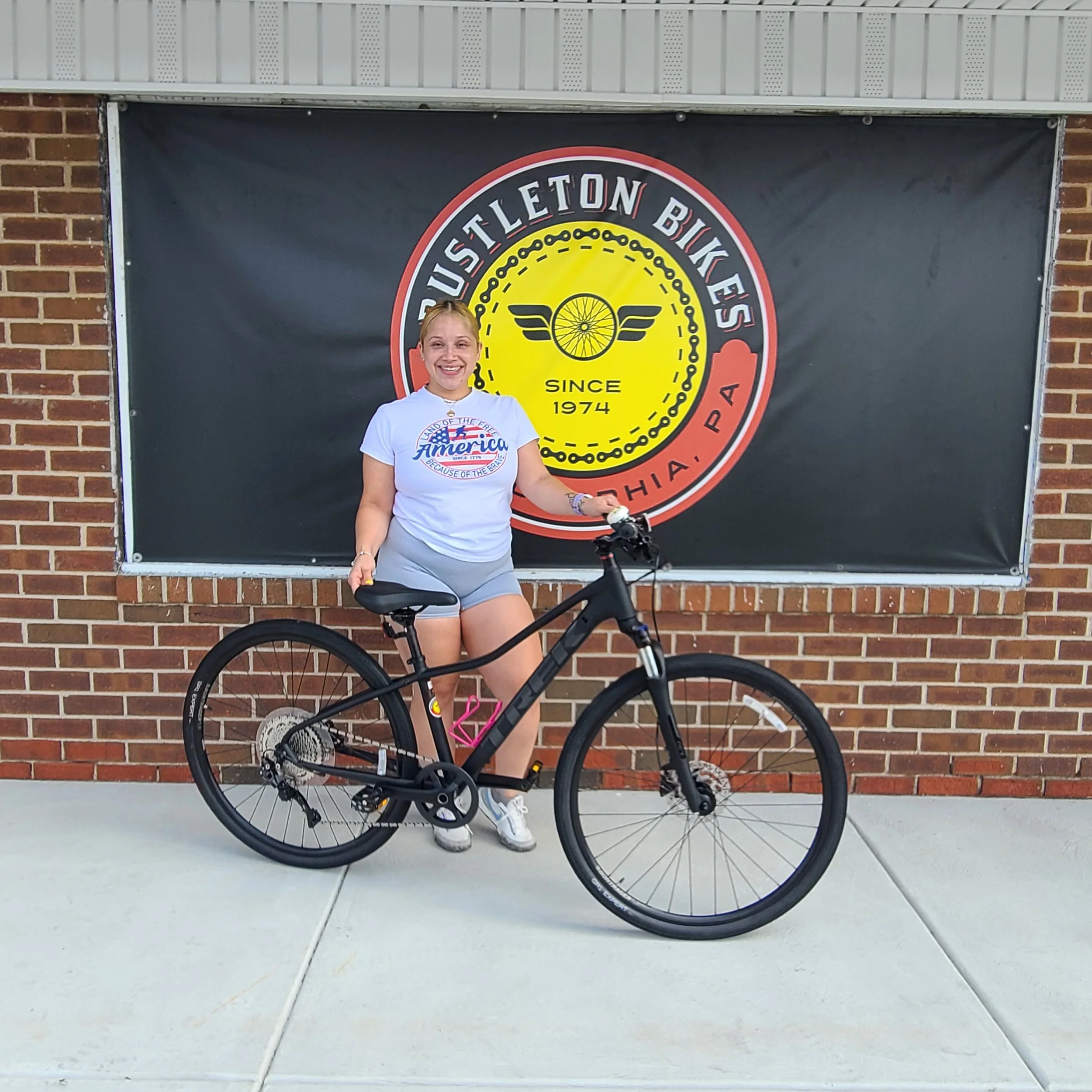 A happy female customer smiles and stands behind her brand-new matte black Trek dual-sport bicycle in front of the shop's brick storefront logo.
