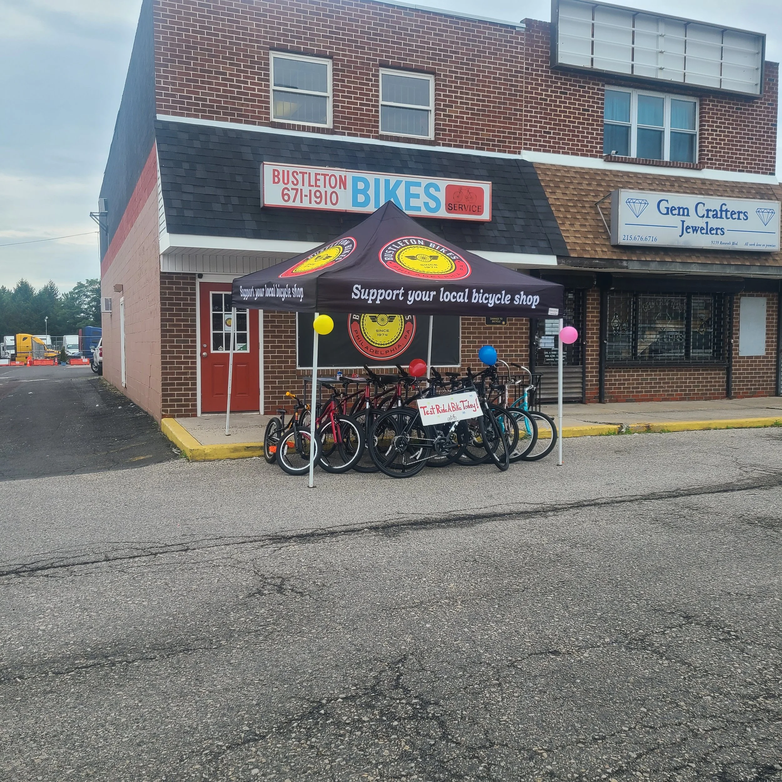 A black canopy tent with the Bustleton Bikes logo reading "Support your local bicycle shop" set up in a parking lot over a row of test-ride bikes.
