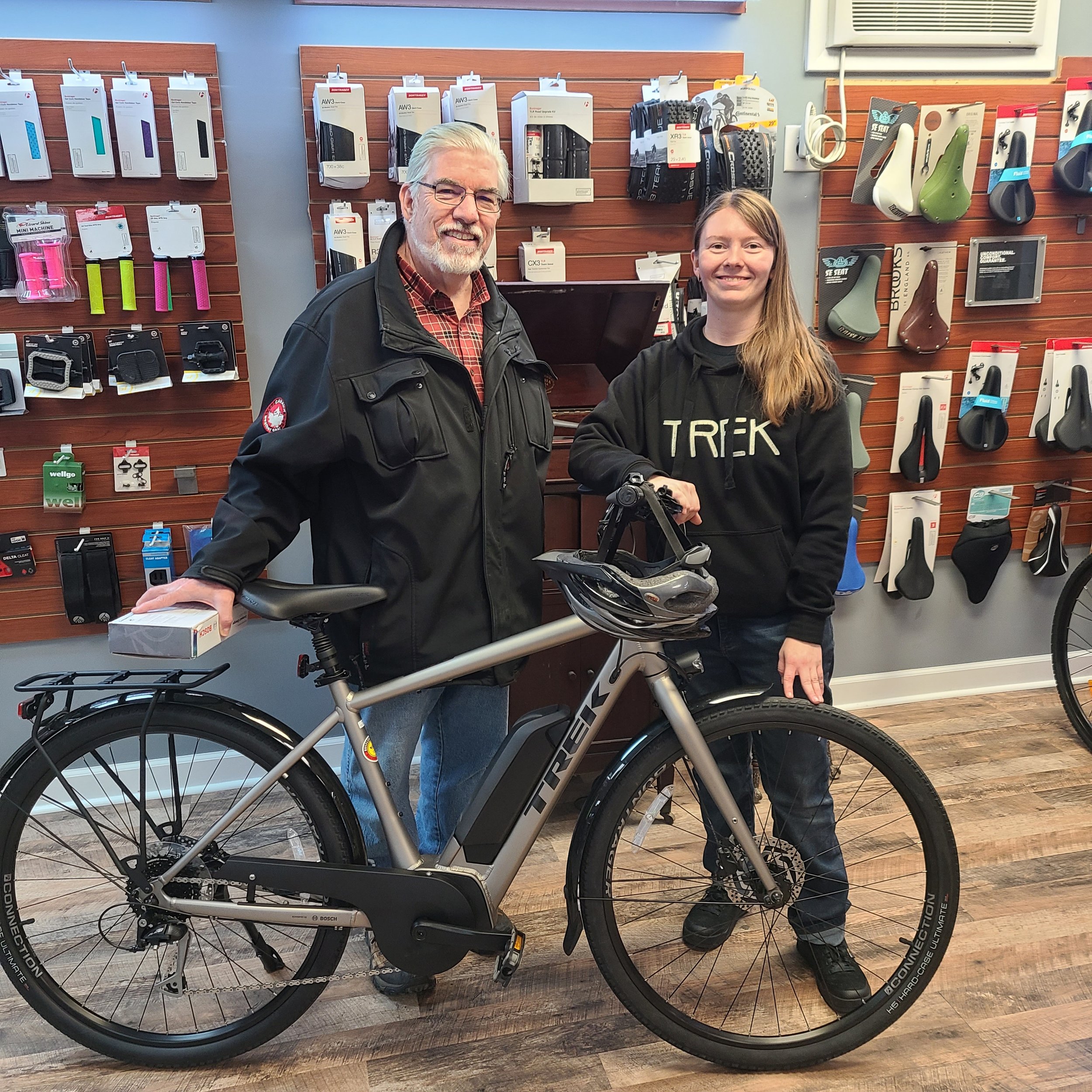 A man and a female bike shop employee smiling inside a bike shop next to a silver Trek Verve+ 2 electric hybrid bicycle.