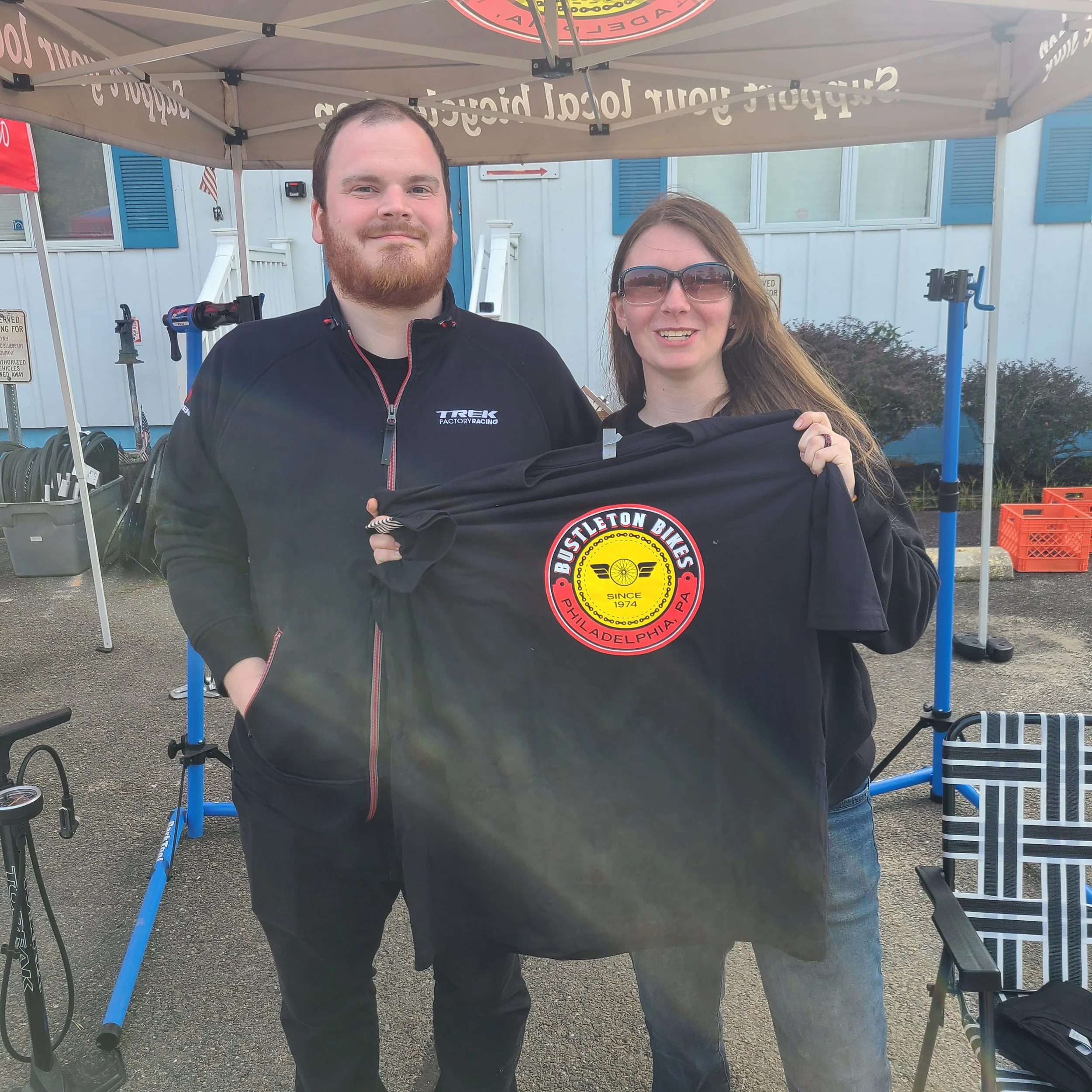 A man in a Trek Factory Racing jacket and a woman holding up a black Bustleton Bikes t-shirt under a shop canopy tent at an outdoor support event.