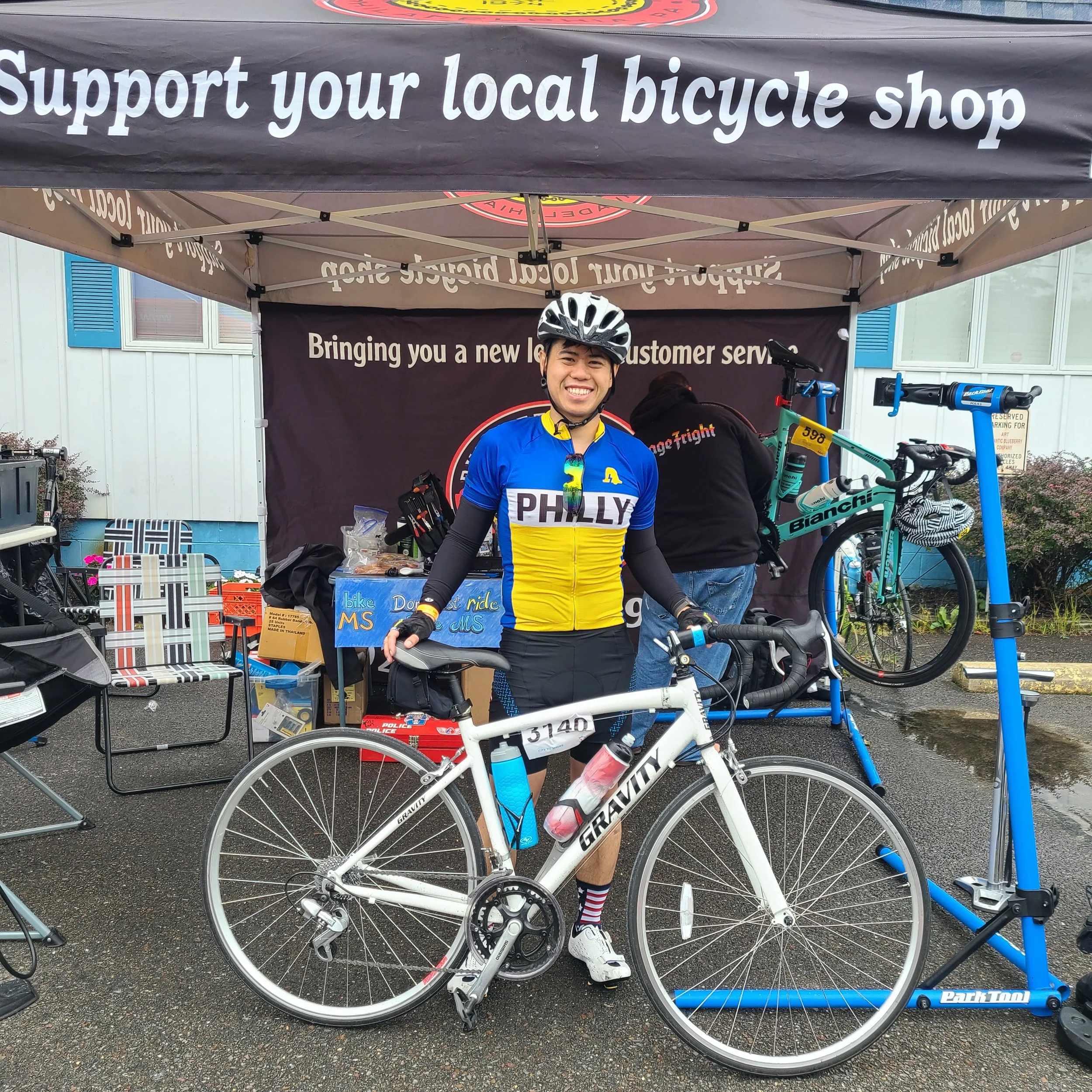A smiling cyclist named Ed wears a "Philly" jersey and stands with his white Gravity road bike under the Bustleton Bikes mechanical support tent at the MS Bike Ride.